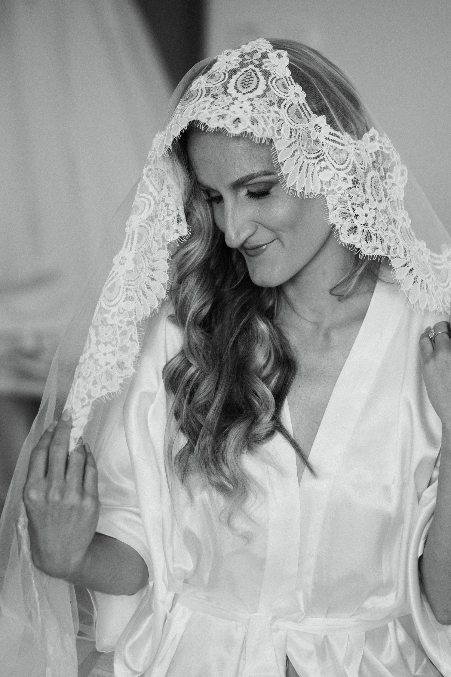 Black and white portrait of a smiling bride wearing a lace mantilla veil and white satin robe with wavy hair.