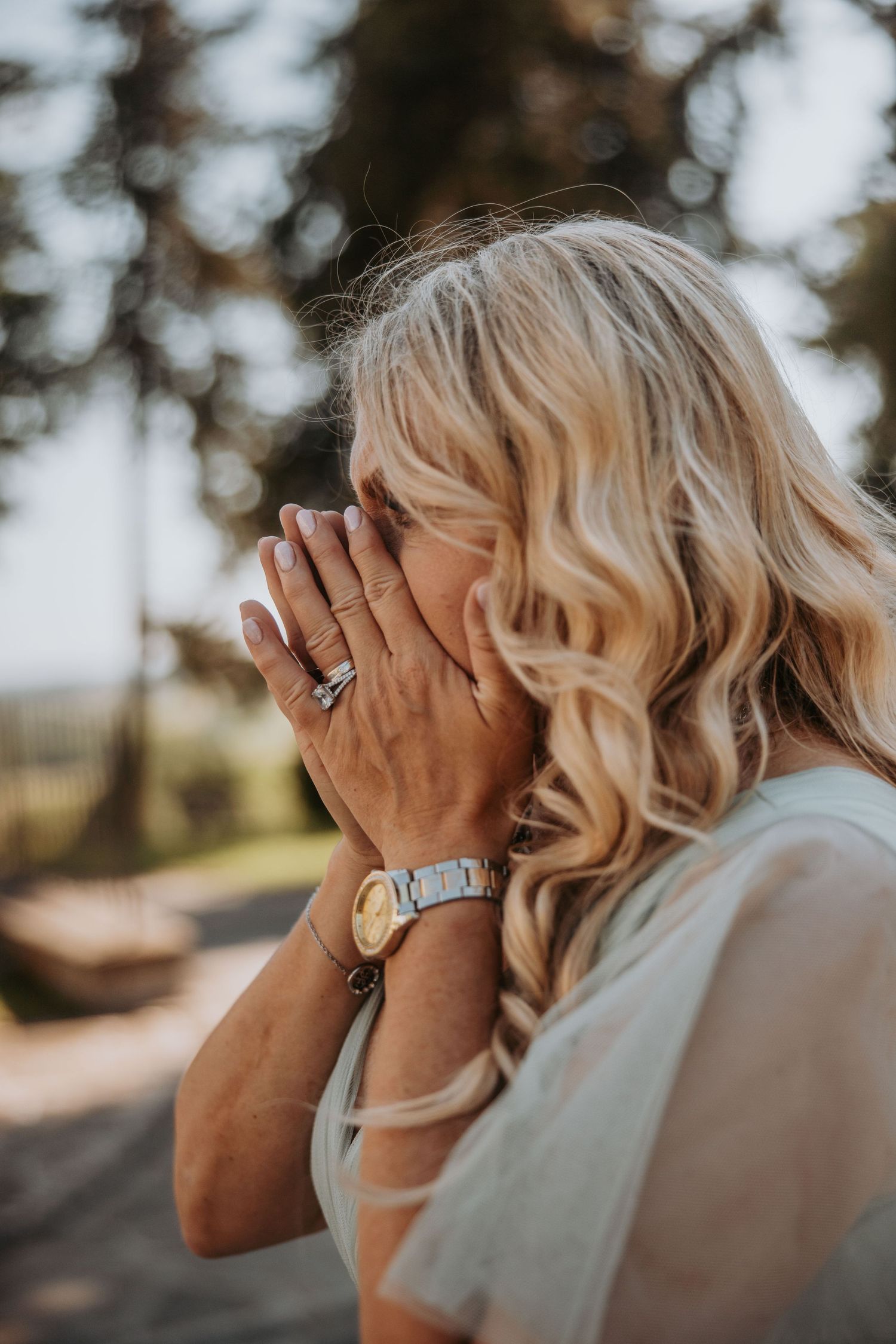 Blonde woman with wavy hair covering her face with hands, wearing a watch and rings outdoors.
