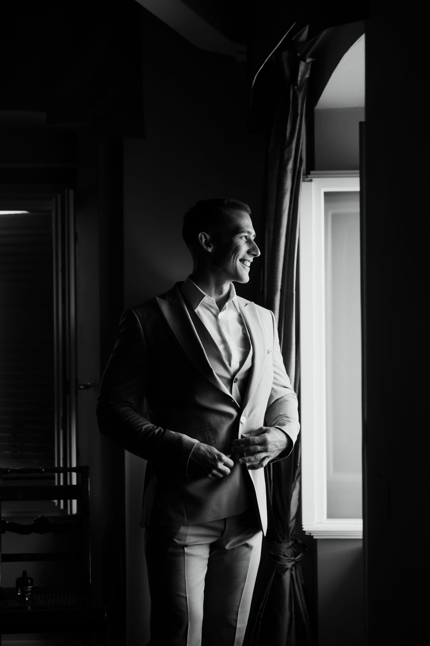 Groom in beige suit smiling by window in black and white wedding photo.