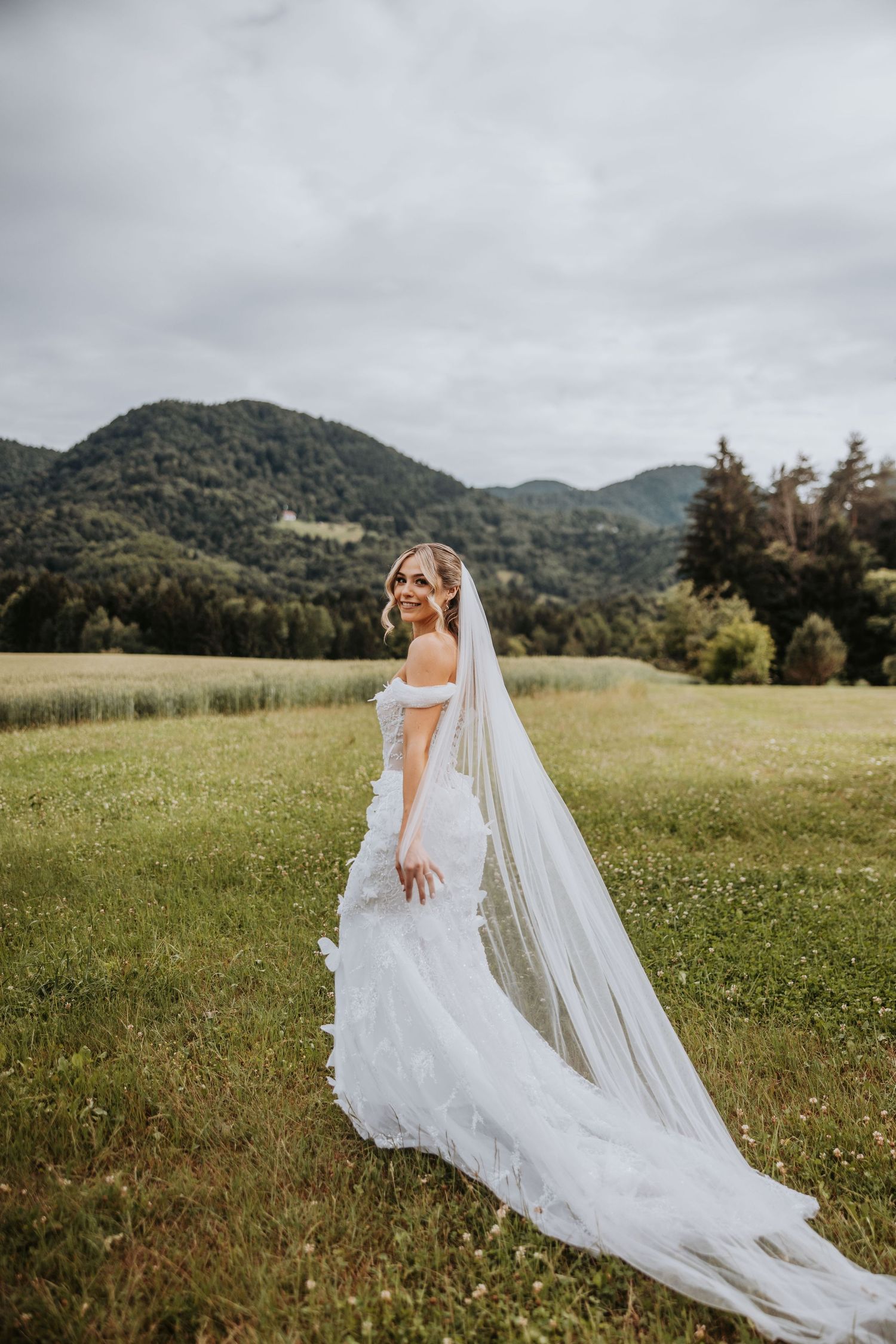 Bride in off-shoulder lace gown with long cathedral veil posing in a green meadow with mountains.