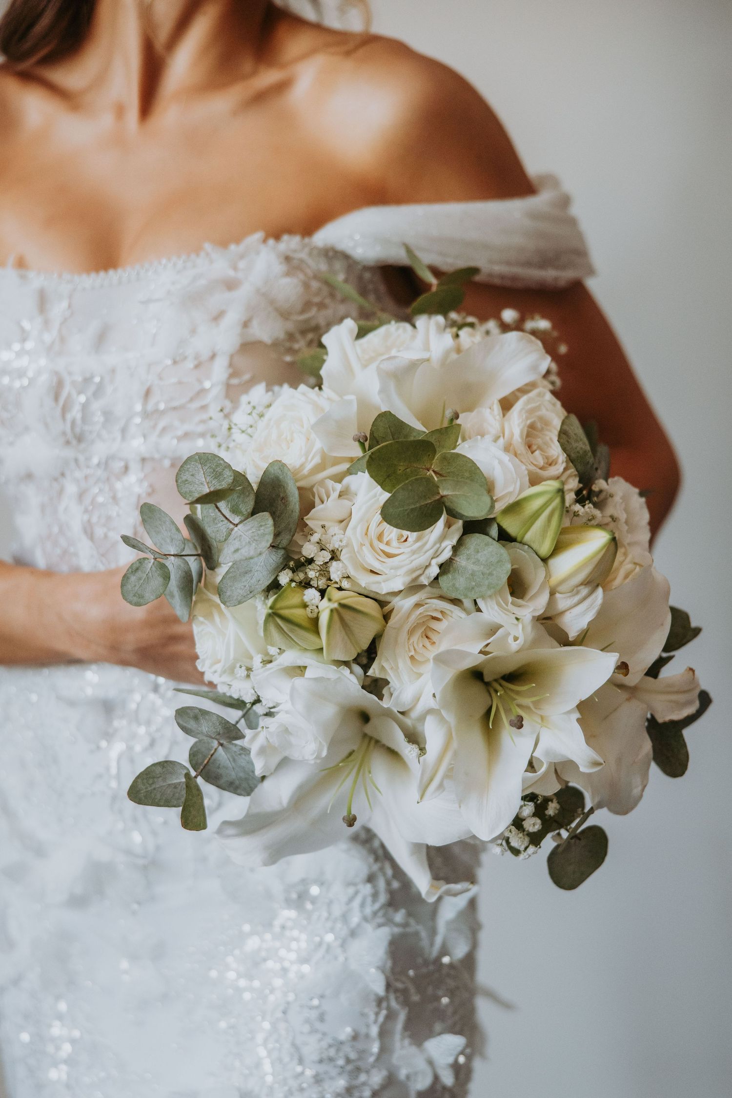 Bride holding elegant white bouquet of lilies, roses, and eucalyptus on her wedding day.