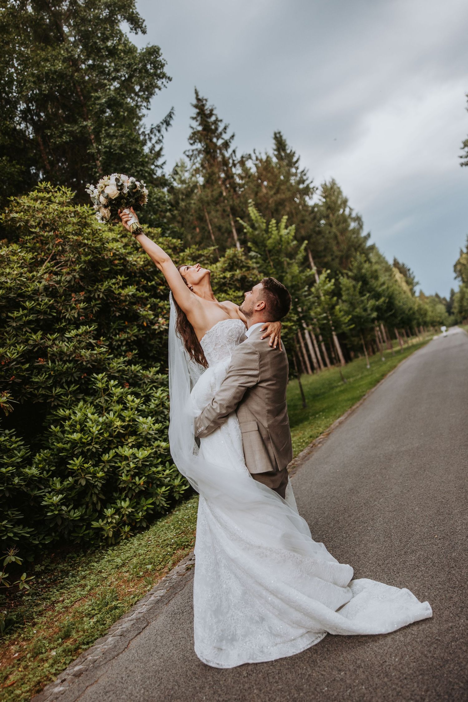 Bride in white gown holds bouquet aloft as groom lifts her on a tree-lined road during outdoor wedding photos.