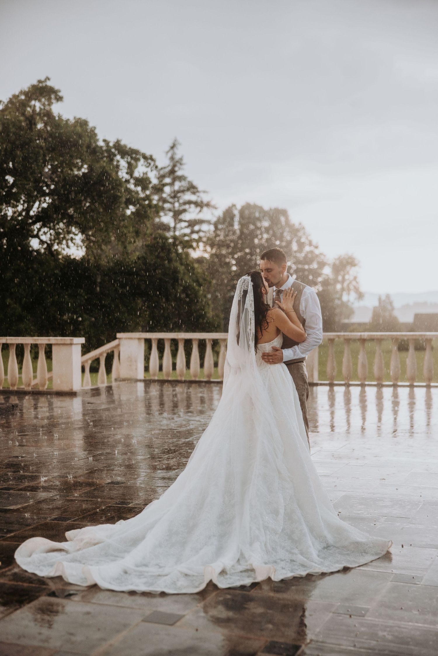 Bride and groom embracing in the rain on a stone terrace in wedding attire.