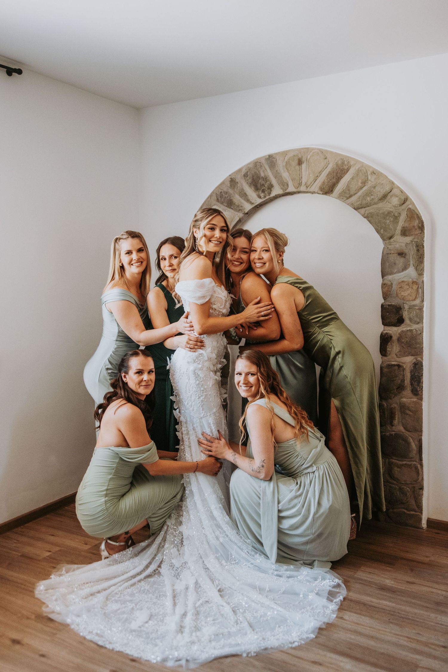 Bride in lace mermaid gown surrounded by bridesmaids in sage green dresses near a stone arch indoors.