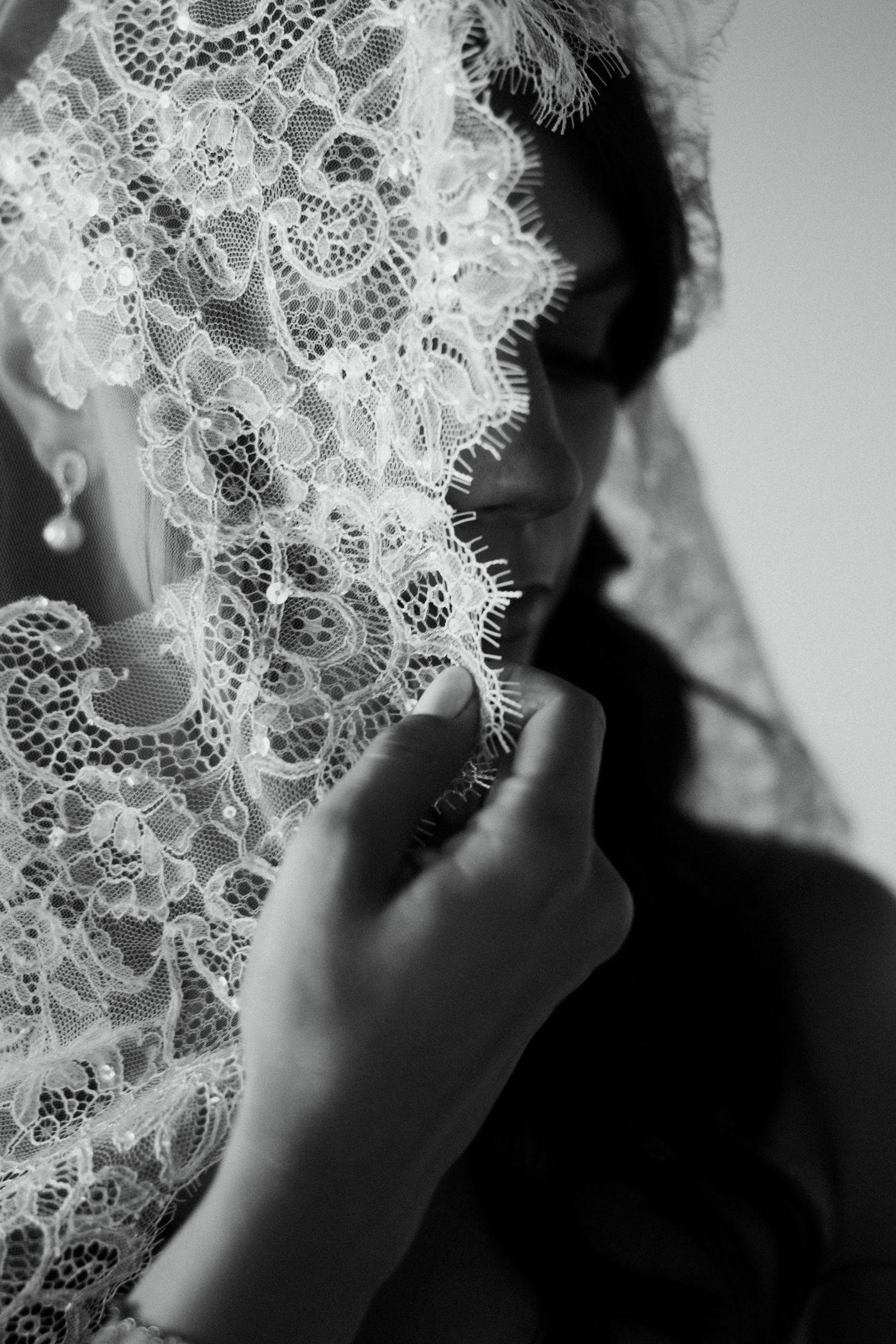 Black and white portrait of a woman holding a delicate lace veil over her face, wearing a pearl earring.