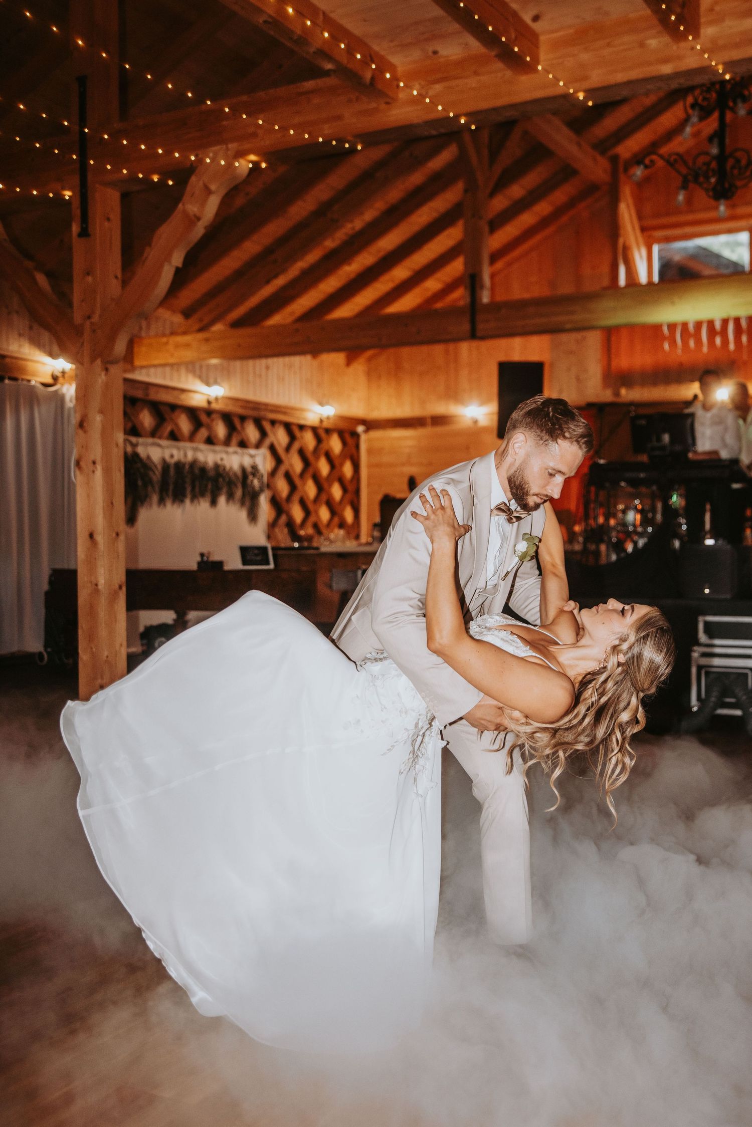 Groom dramatically dipping bride during first dance on foggy dance floor in rustic barn venue.