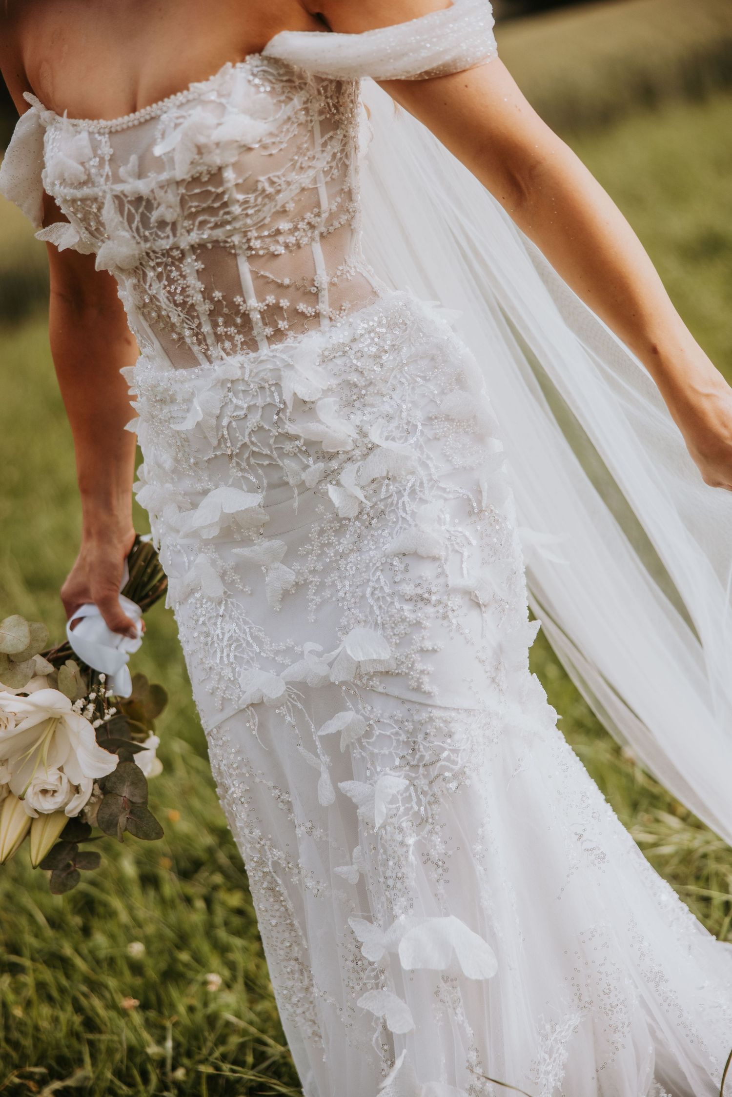 Bride in off-shoulder floral lace wedding gown holding bouquet outdoors.