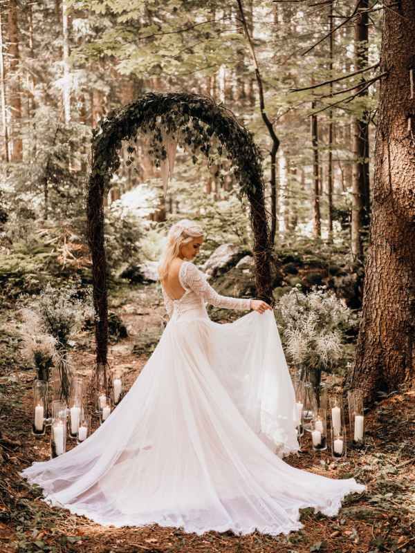 Bride in a flowing white gown stands beneath a floral arch surrounded by candles in an enchanted forest setting.