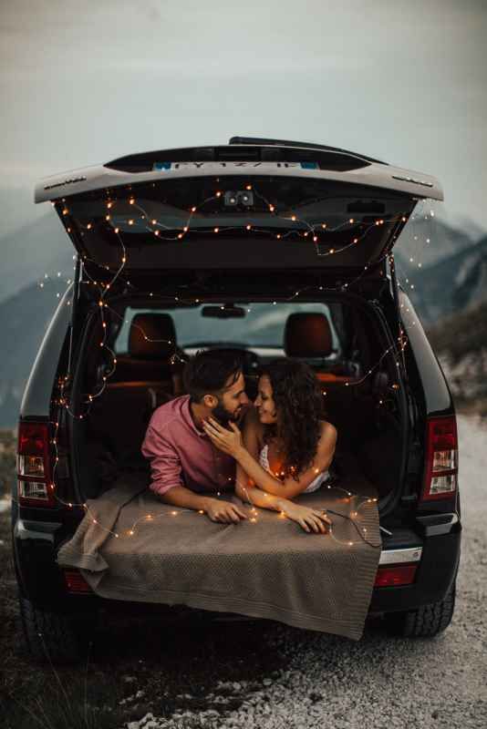 Romantic couple lying in car trunk decorated with fairy lights, sharing intimate moment with mountain backdrop.