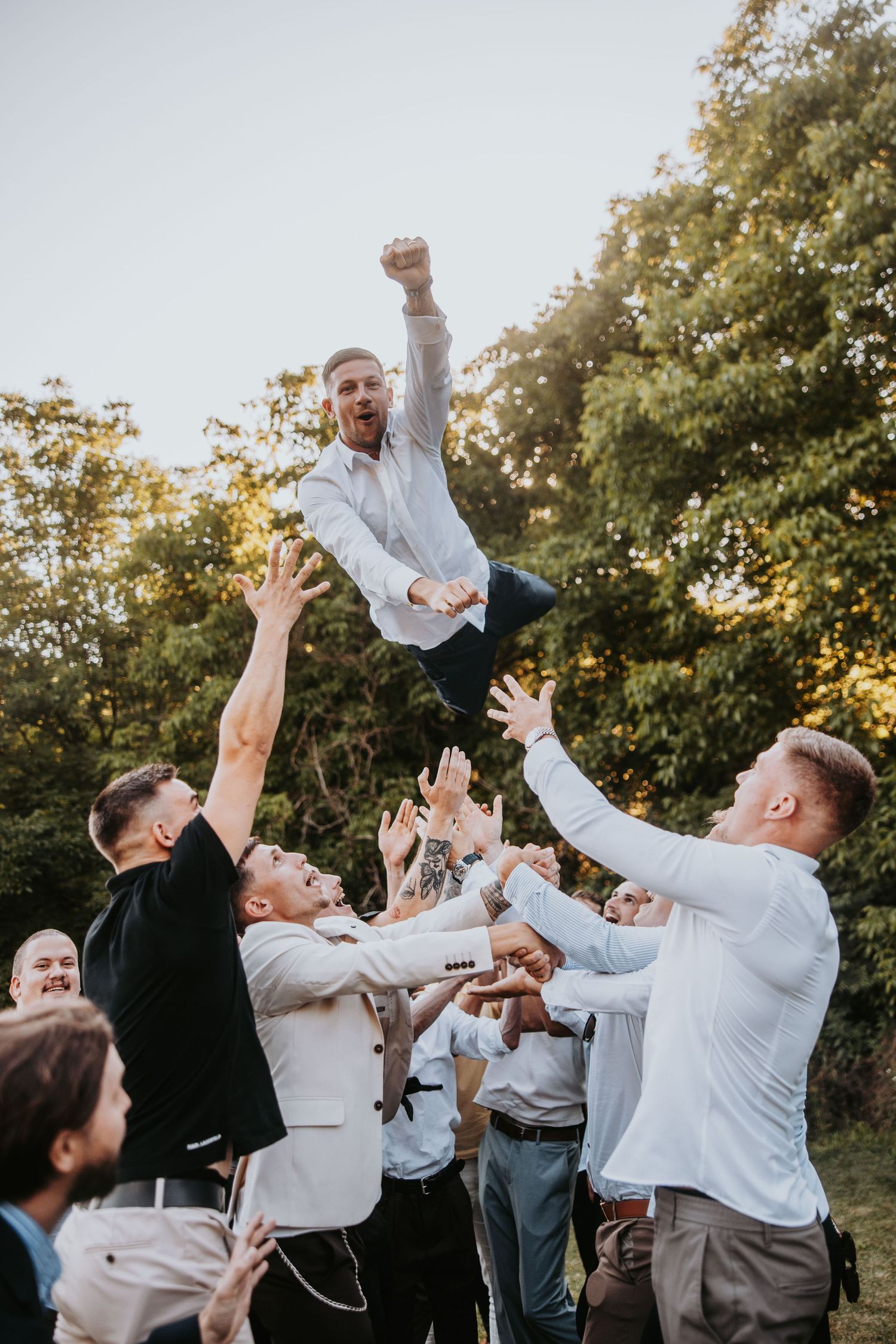 Groomsmen tossing groom into the air in celebration at outdoor wedding reception near trees.