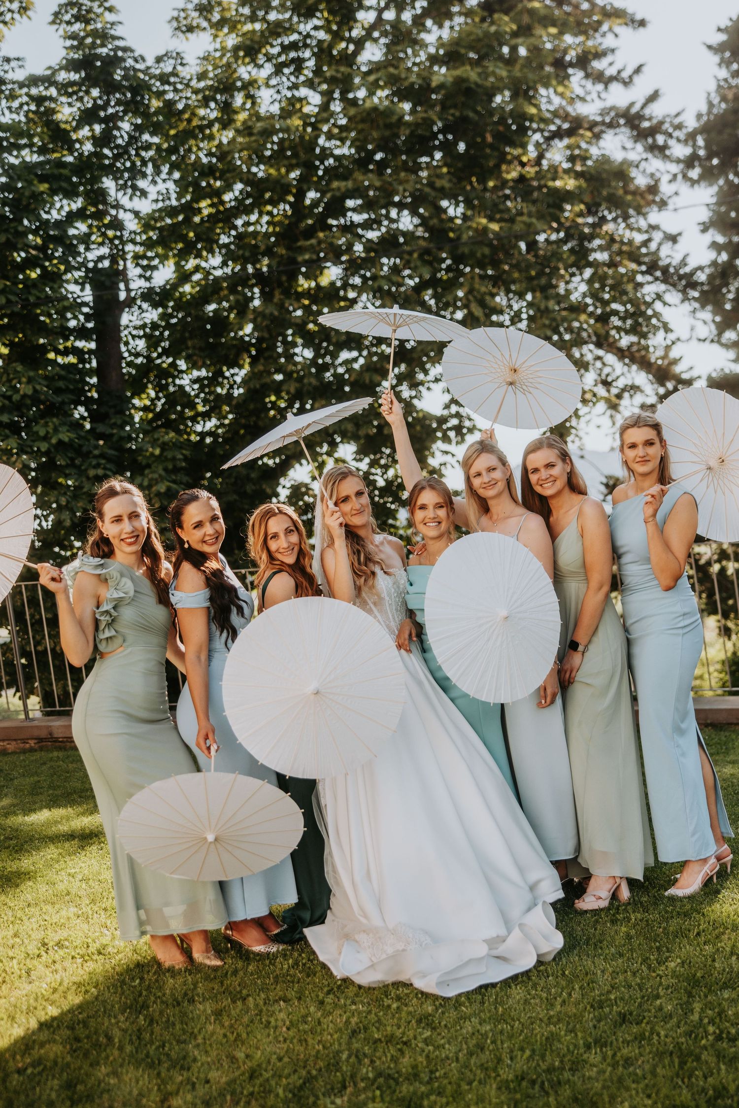 Bride and seven bridesmaids in sage green dresses posing joyfully with white parasols in garden.