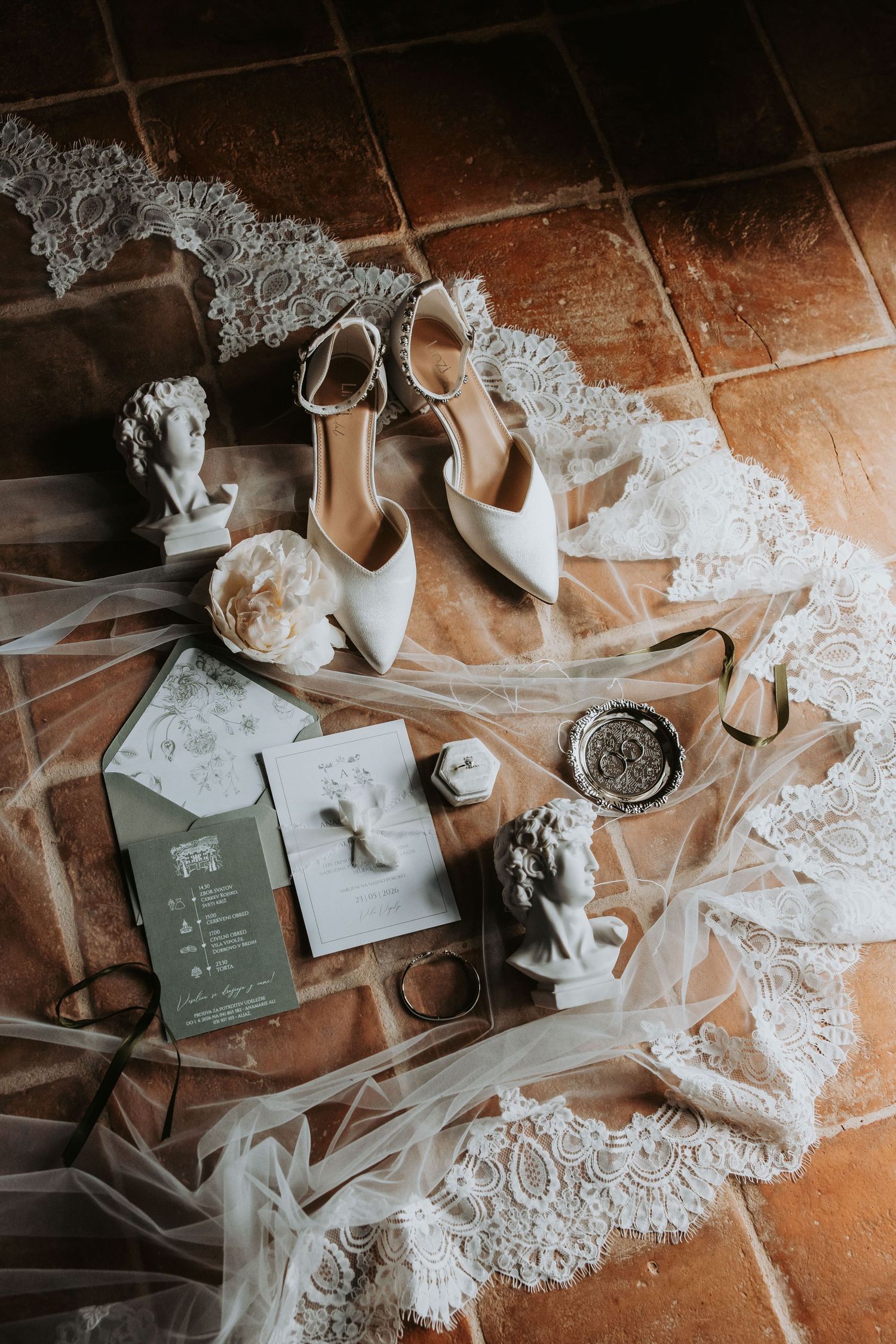 Elegant bridal flat lay with white heels, lace veil, wedding invitations, and decorative accessories on terracotta tiles.