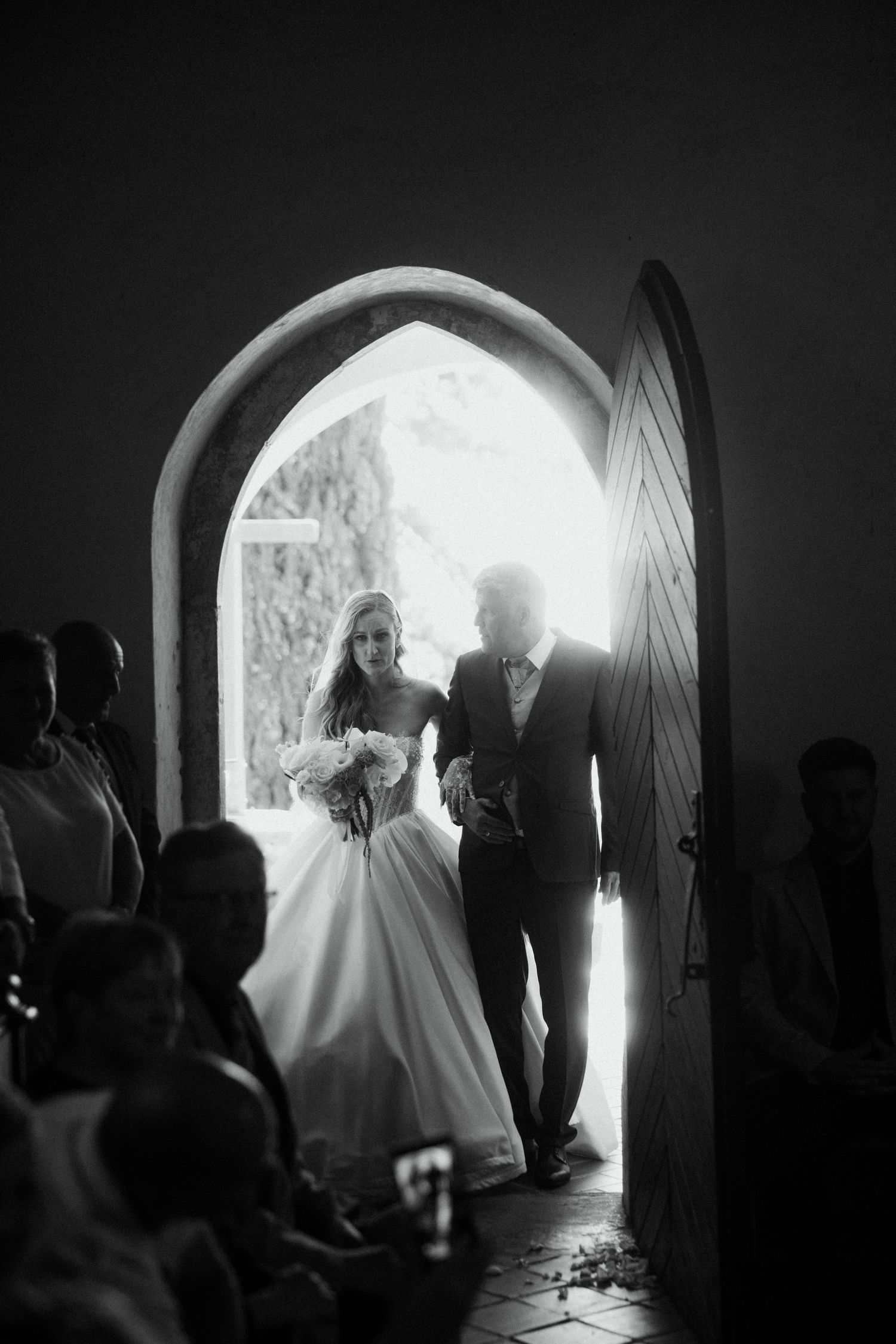 Bride in white gown enters church through arched doorway escorted by father in black and white photo.