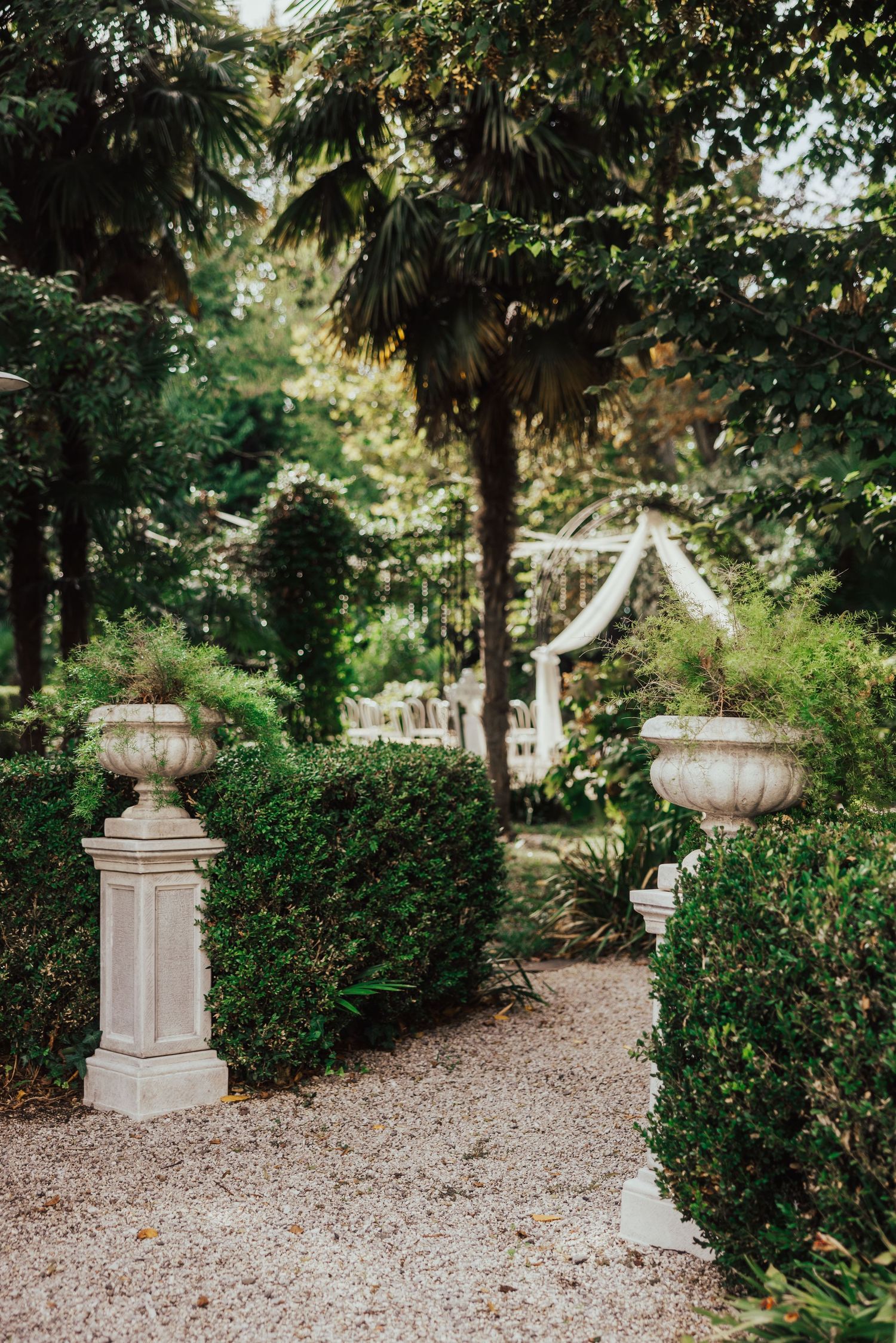Lush garden wedding venue pathway with ornate stone urns and a decorated arch visible in the background.