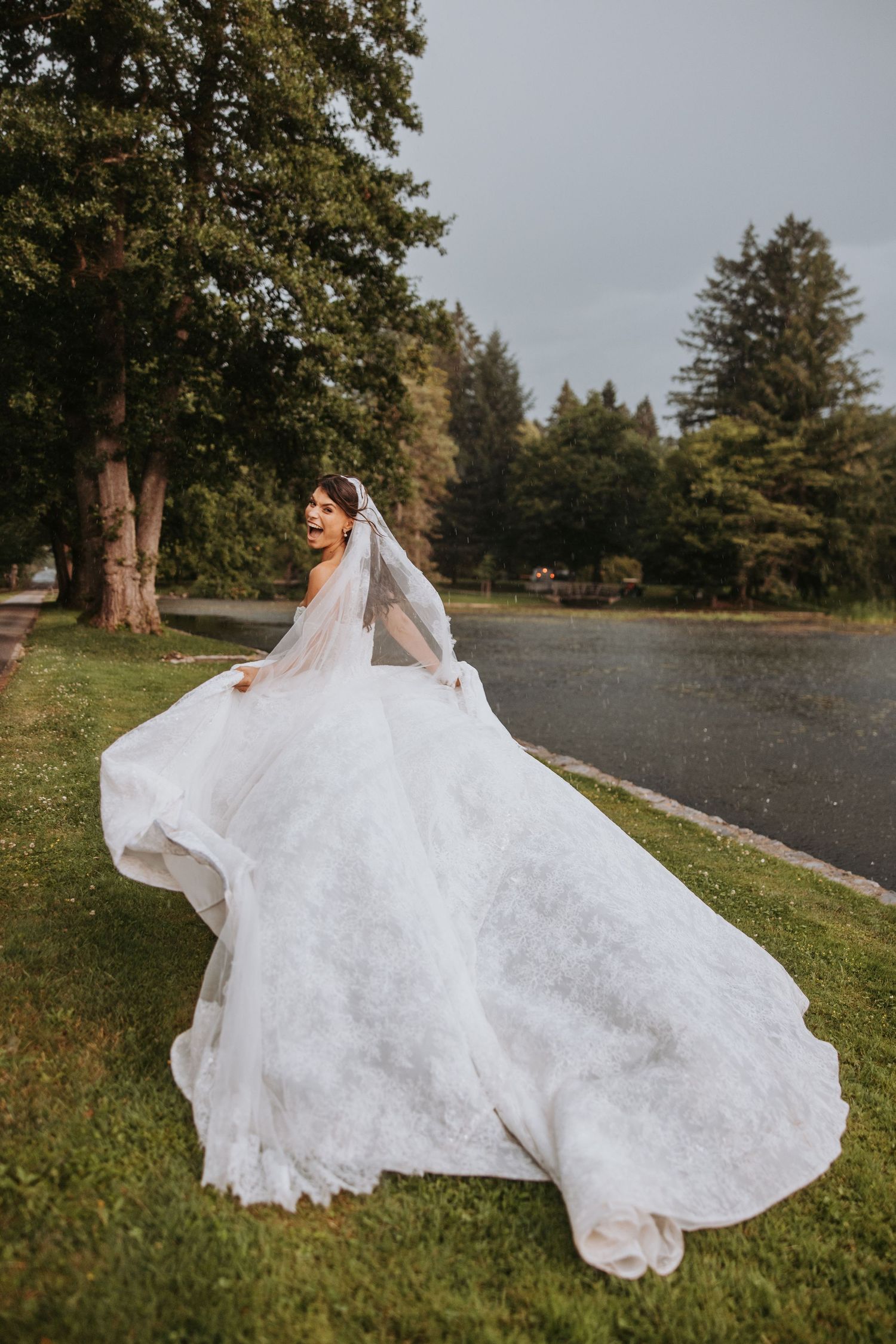 Bride in flowing white lace gown and veil twirls joyfully outdoors near a lake surrounded by trees.