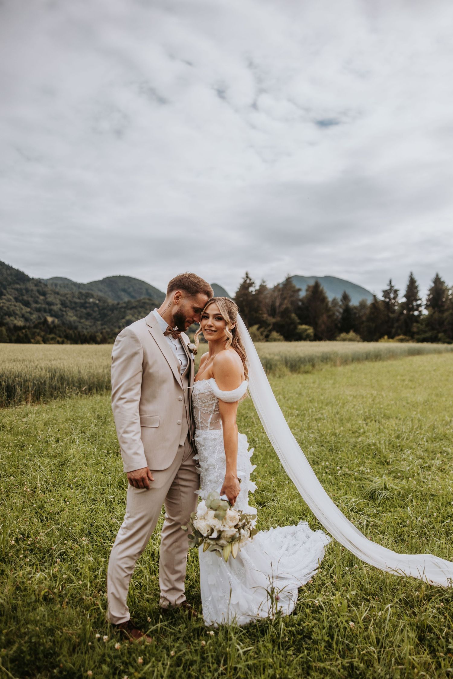 Bride and groom pose in lush green field with flowing veil and mountain scenery under cloudy skies.