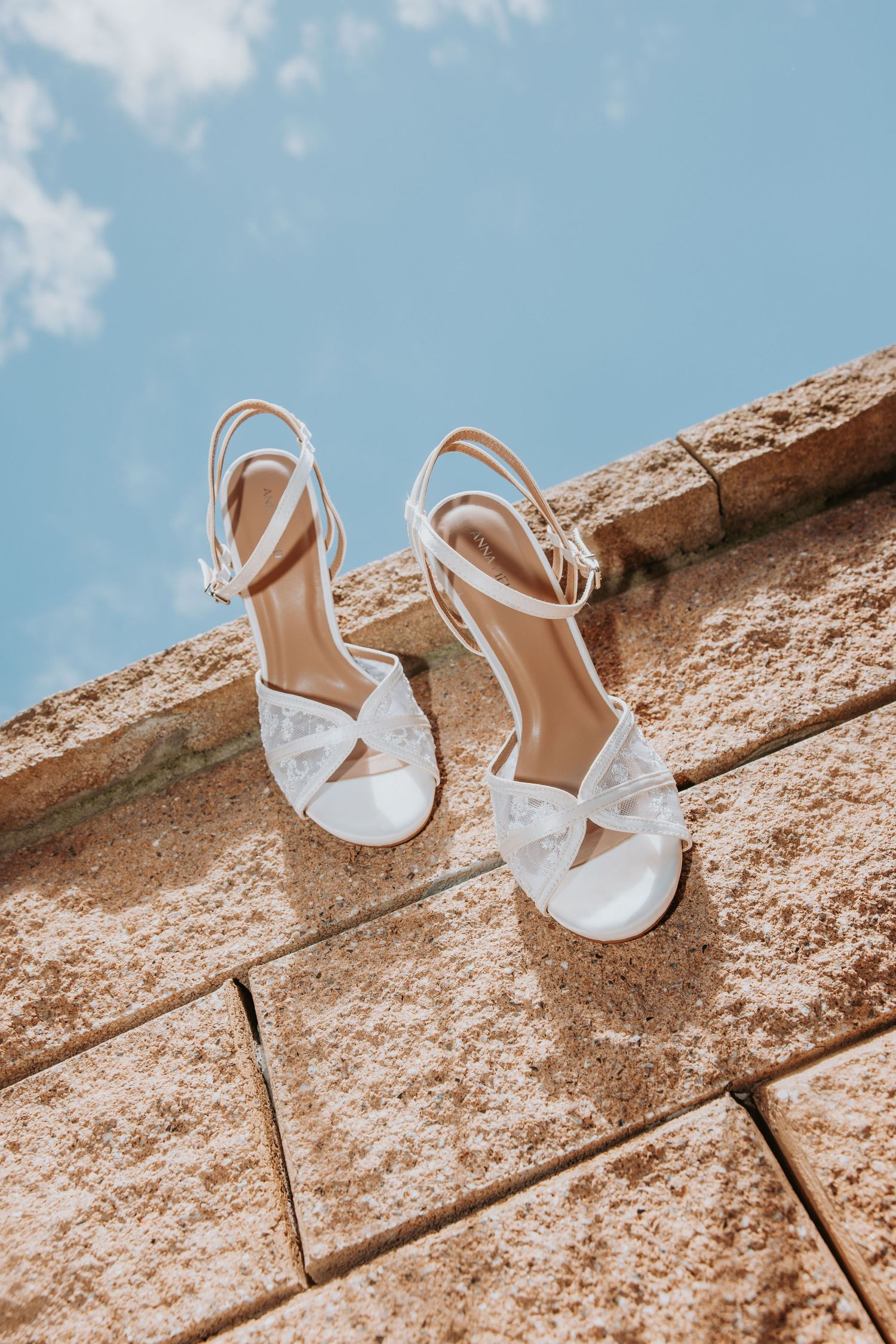 White bridal heeled sandals with ankle straps resting on a stone wall against a blue sky.