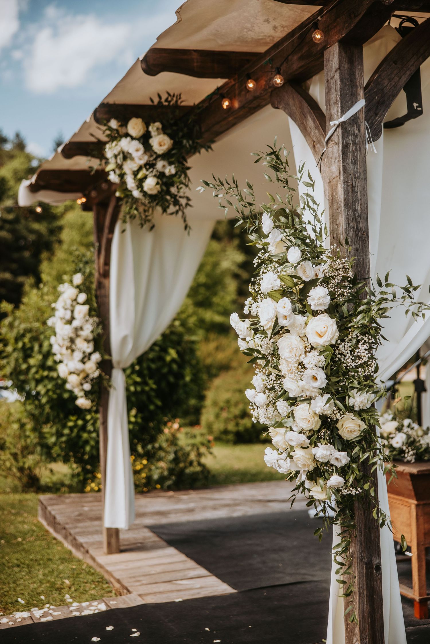 Rustic wooden wedding arch adorned with white roses and greenery, with white draping curtains outdoors.