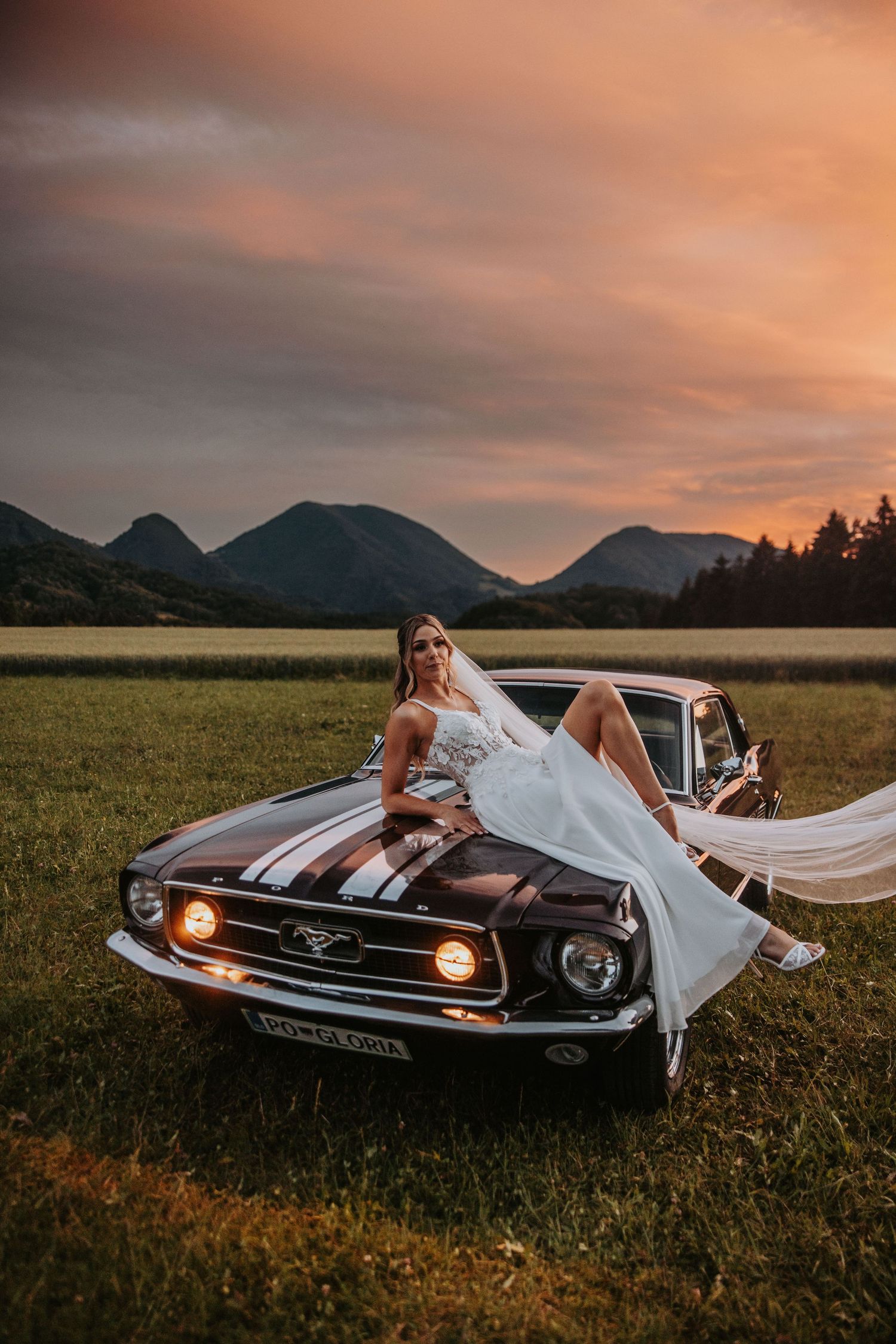 Bride in white gown and veil posing on a classic Ford Mustang at sunset with mountain backdrop.