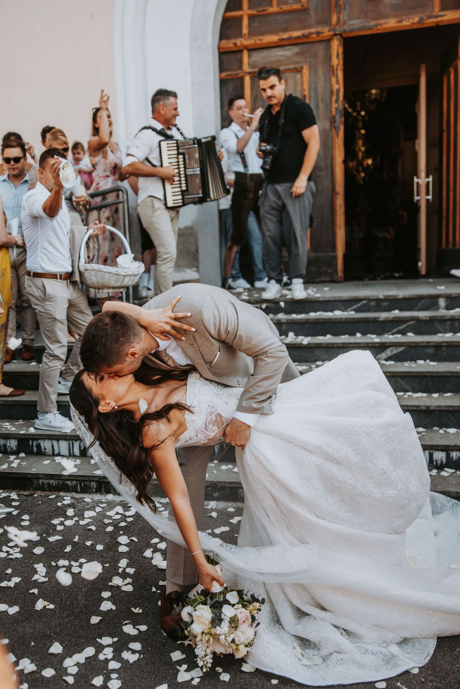 Bride and groom share a romantic dip kiss on church steps surrounded by wedding guests and accordion player.