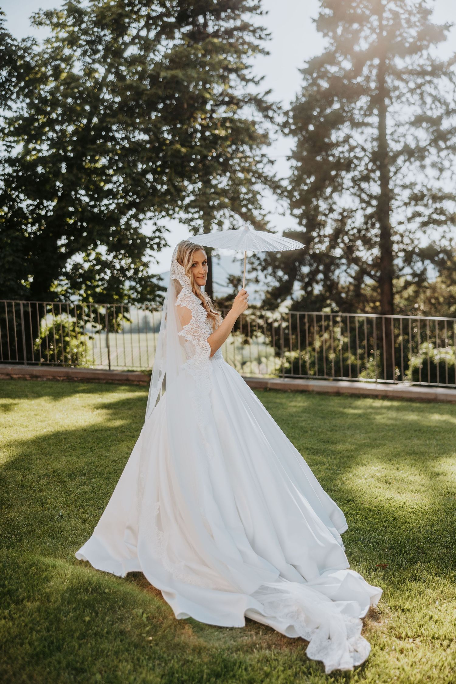 Bride in elegant white ball gown with lace back and veil poses outdoors in sunlit garden.