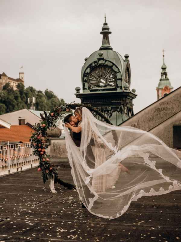 Bride and groom kissing under floral arch, veil flowing in wind with historic clock tower backdrop.