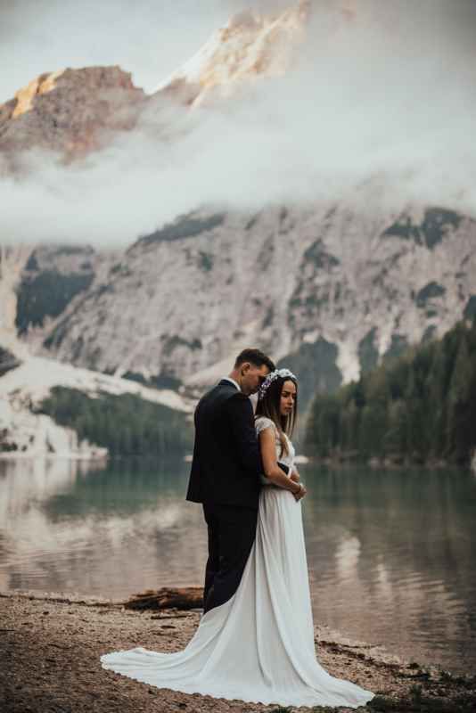 Bride and groom embracing by a mountain lake with misty peaks and forest backdrop on their wedding day.
