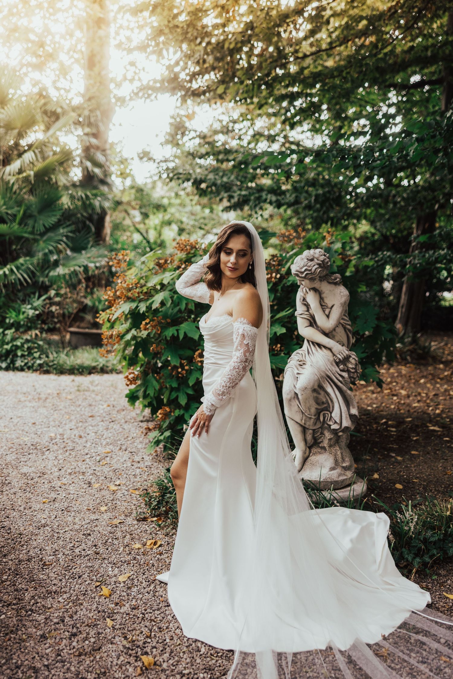 Bride in an off-shoulder white gown with lace sleeves poses beside a stone angel statue in a sunlit garden.