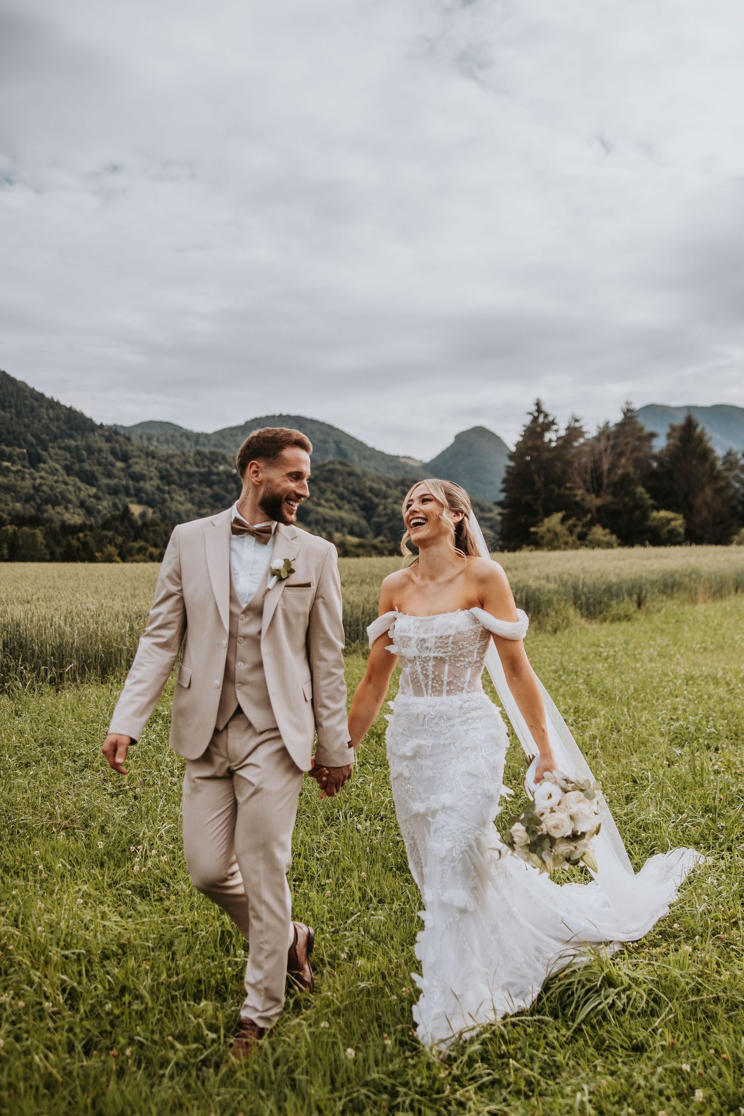 Happy bride and groom walking hand in hand through green meadow with mountain backdrop.