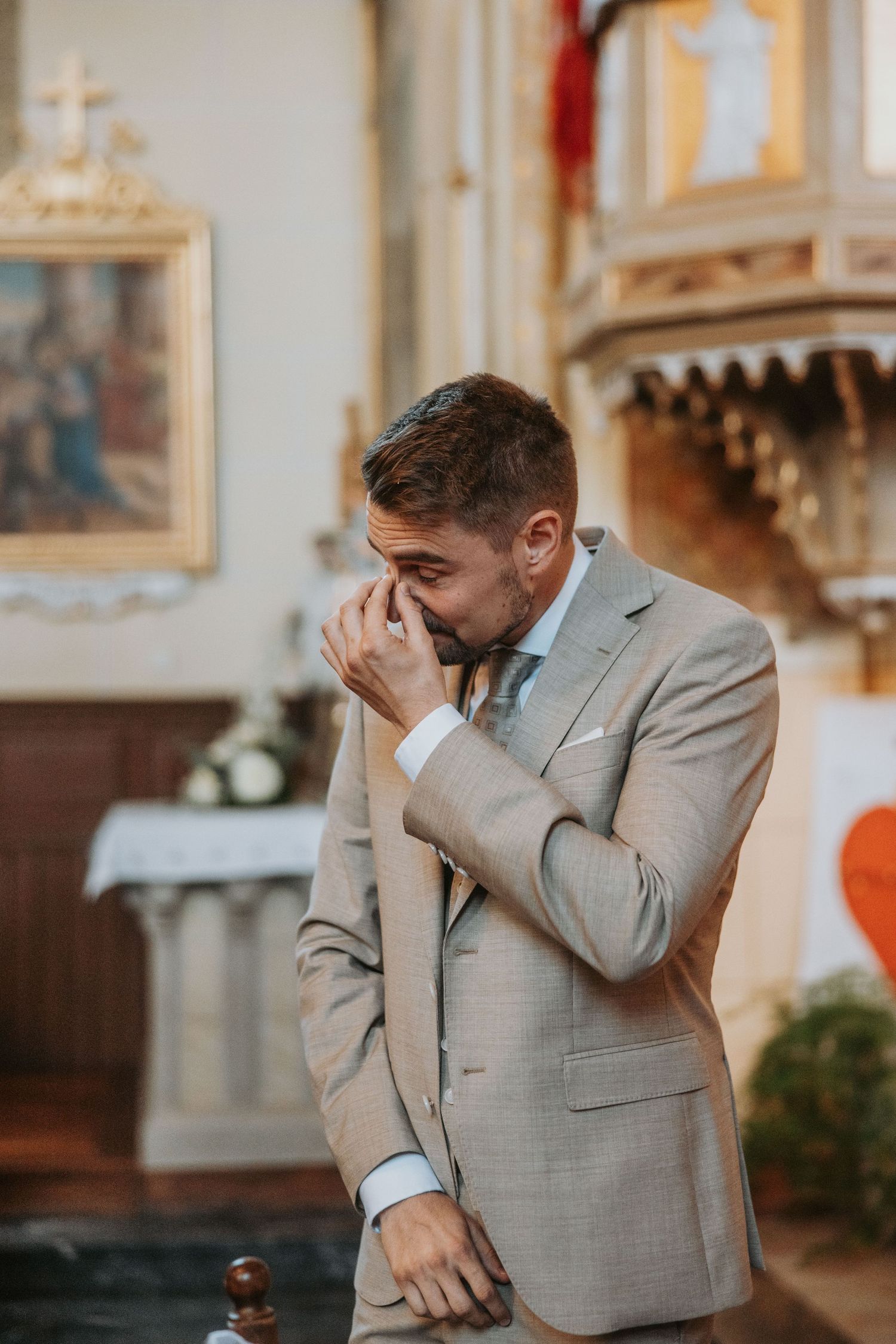 Emotional groom in beige suit covers his mouth with hands while waiting at the church altar.