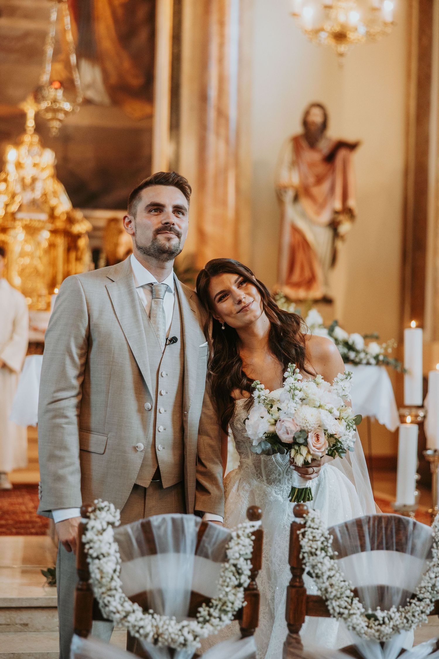 Bride and groom standing together in ornate church interior, bride holding white and pink bouquet, groom in beige suit.