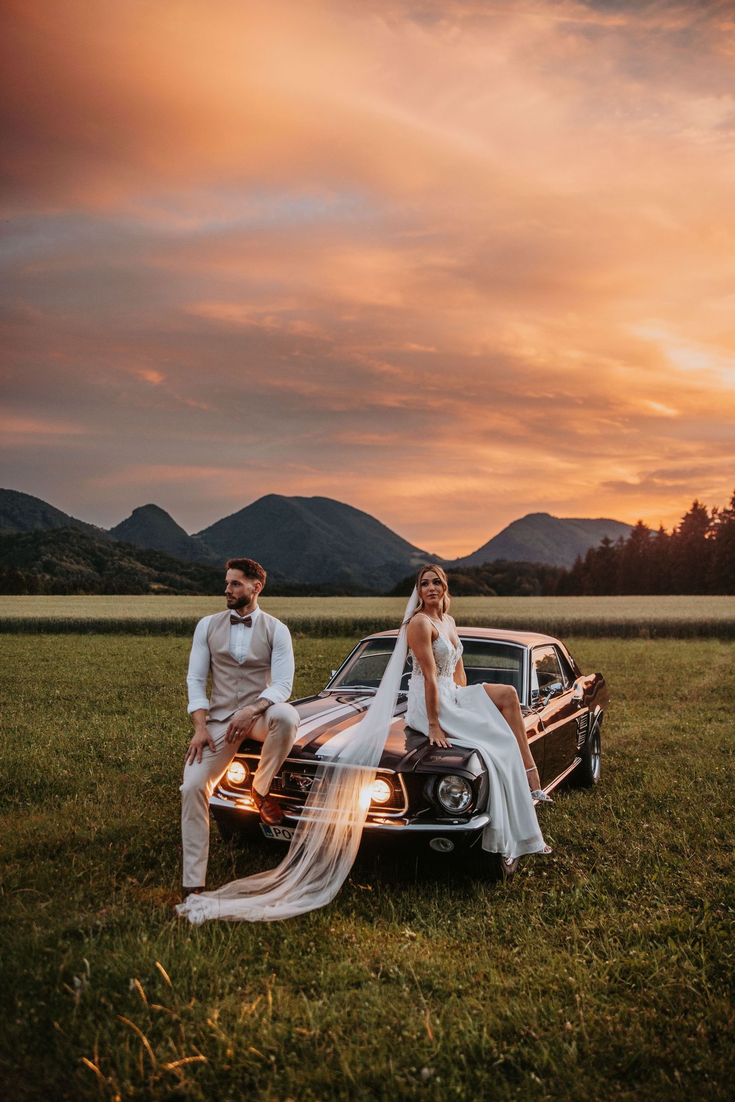 Bride and groom pose on vintage dark Mustang at sunset with dramatic orange sky and mountains.