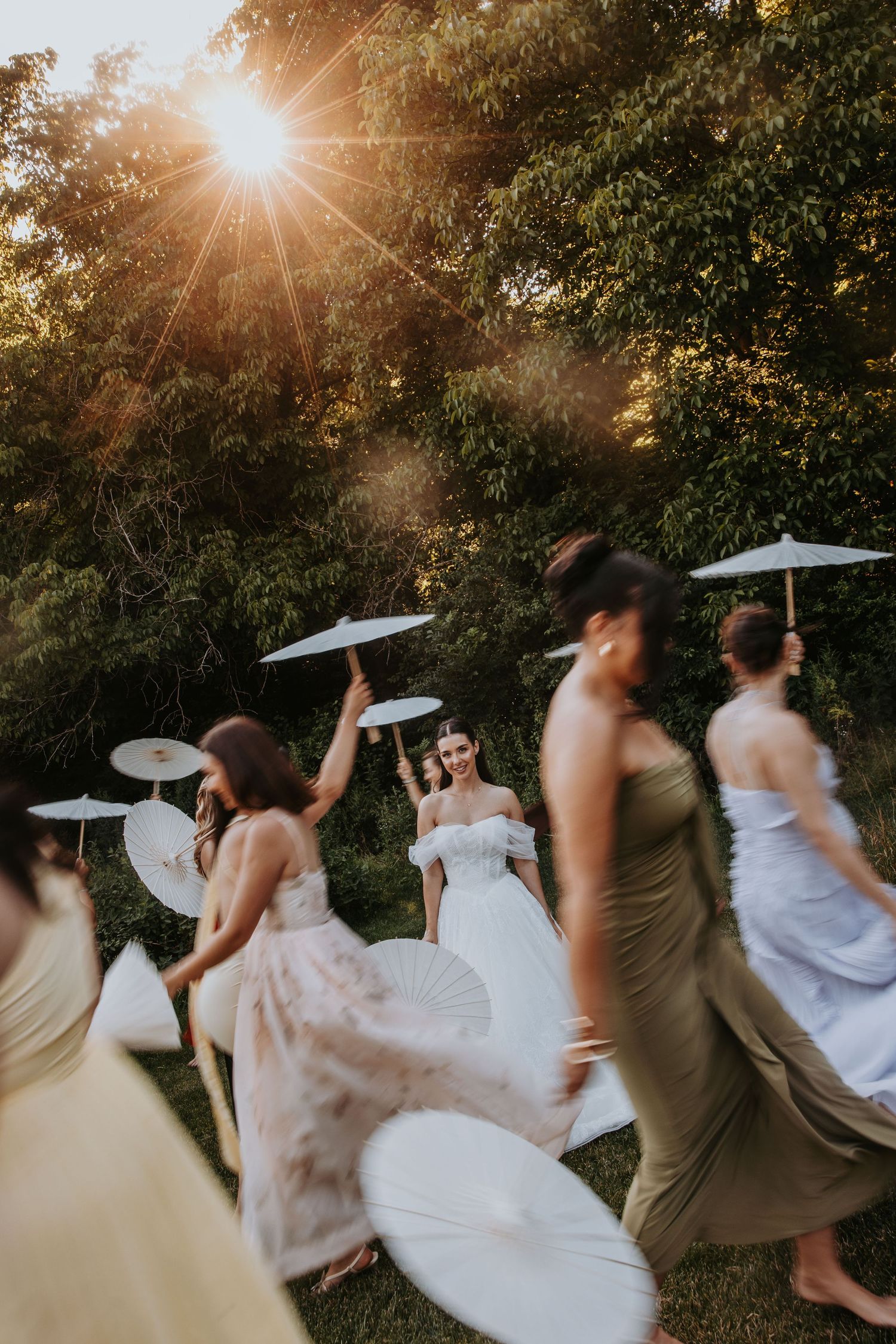 Bridesmaids in mismatched gowns twirl with white parasols in golden sunset light among lush green trees.