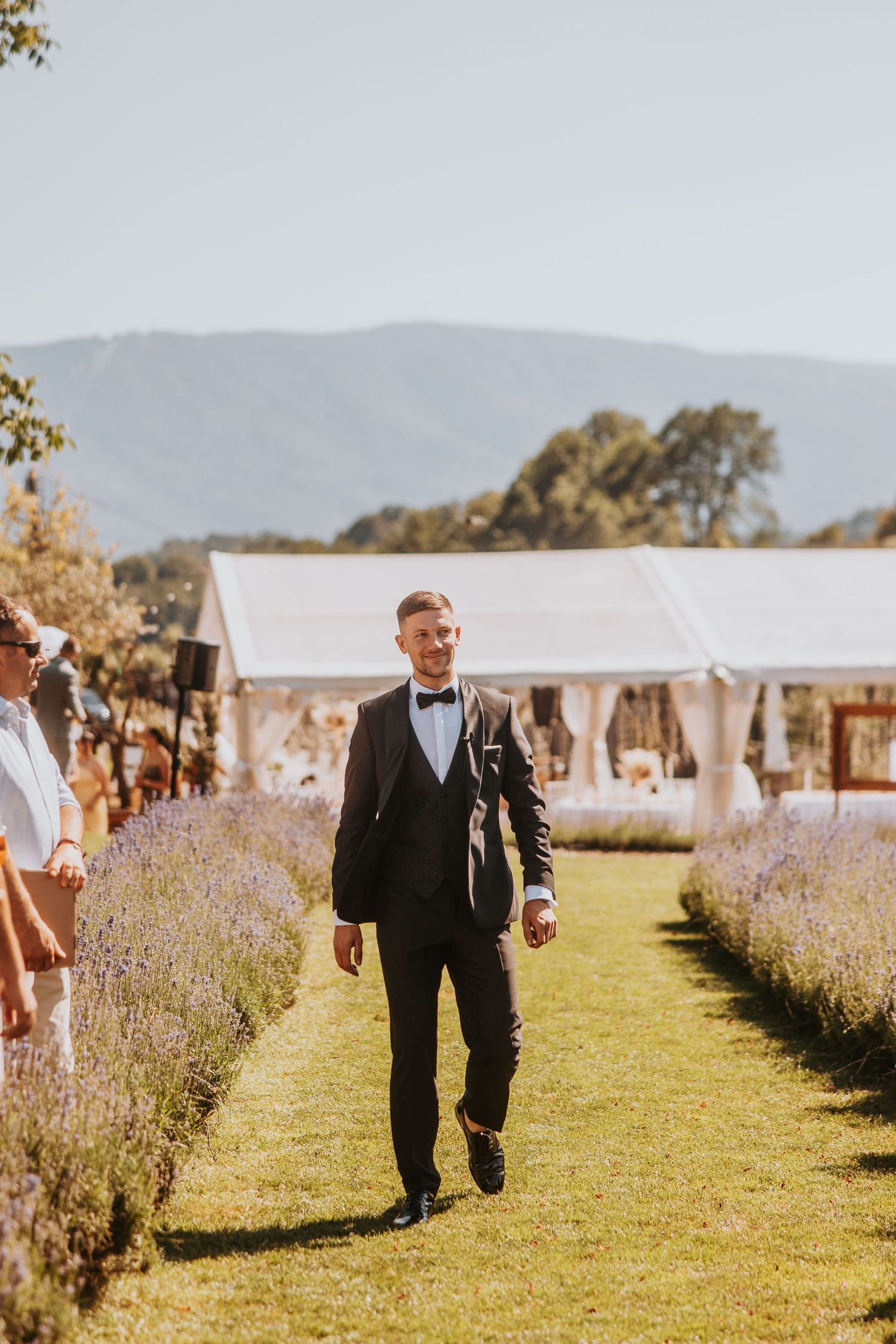 Smiling groom in black tuxedo walking down lavender-lined aisle at outdoor wedding venue with mountain backdrop.