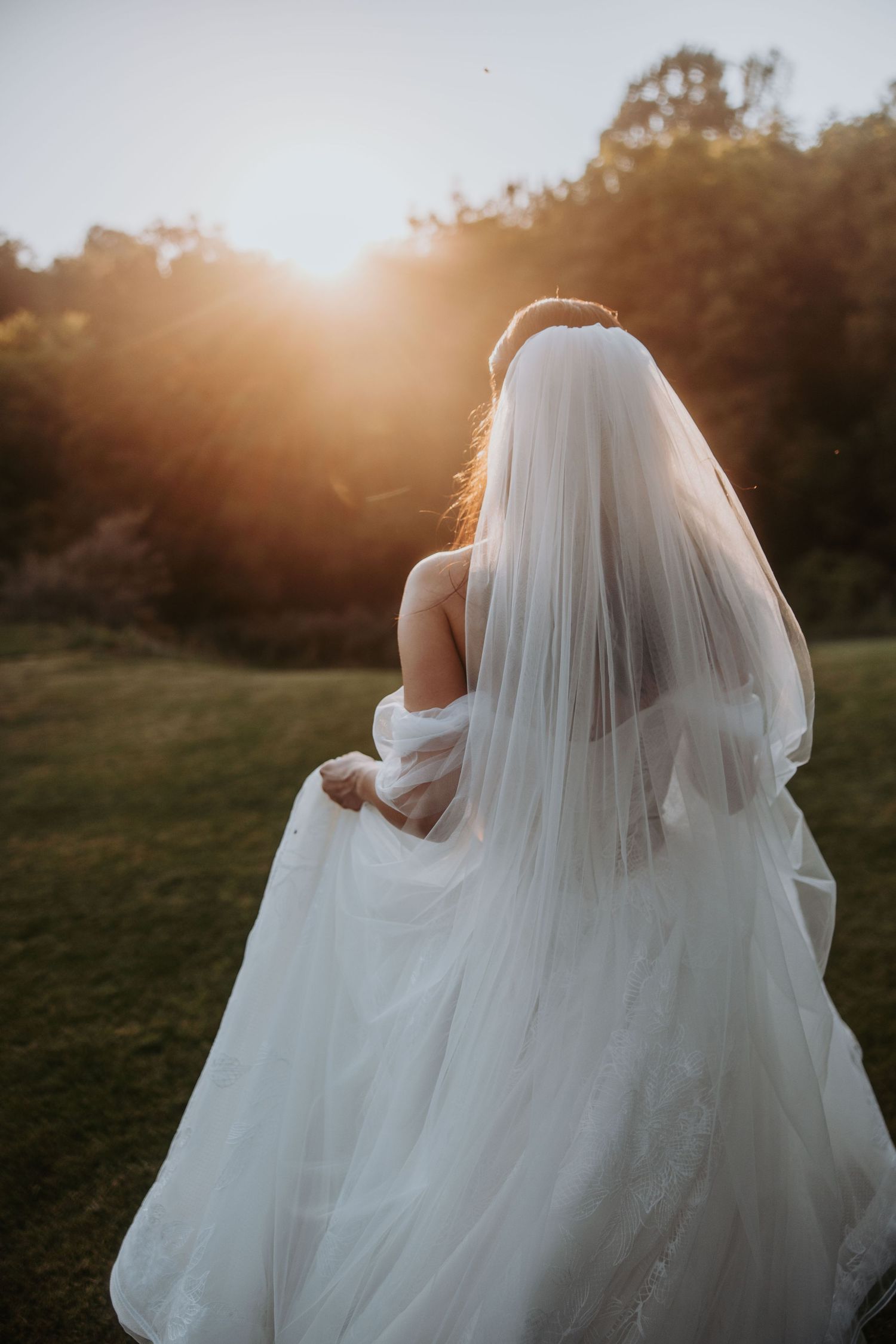Bride in flowing white gown and veil walks toward glowing golden sunset in an outdoor garden setting.