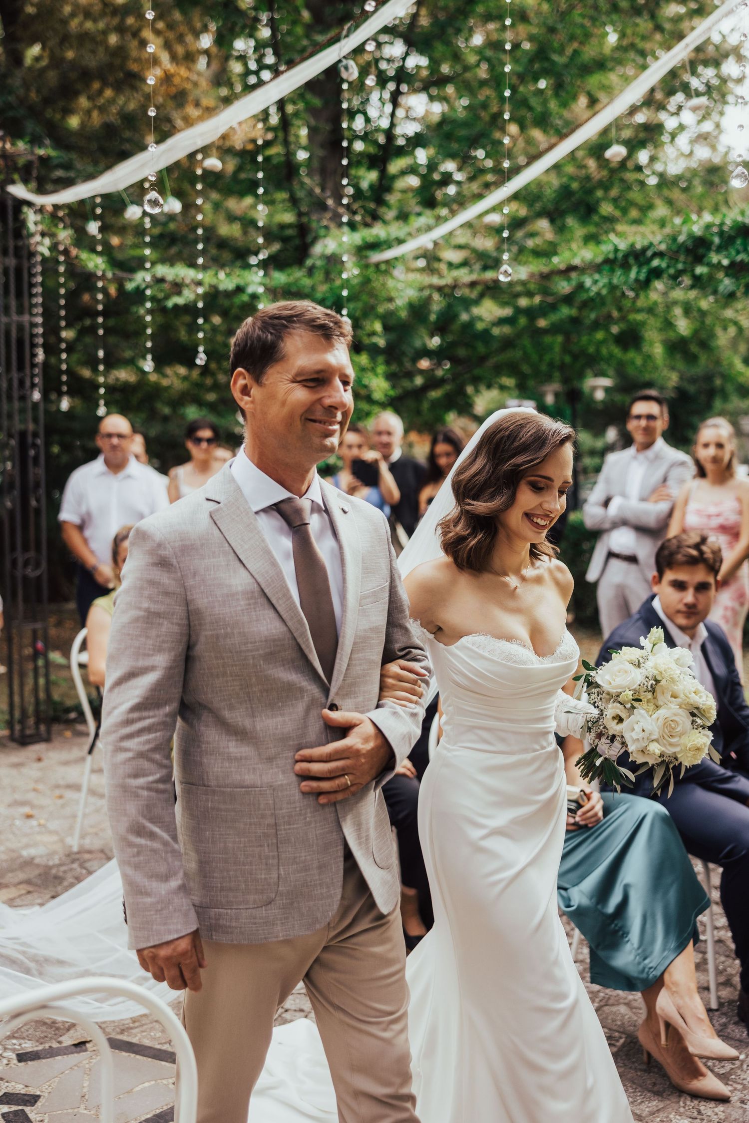 Smiling bride in strapless gown and escort walk down aisle at outdoor garden wedding ceremony.