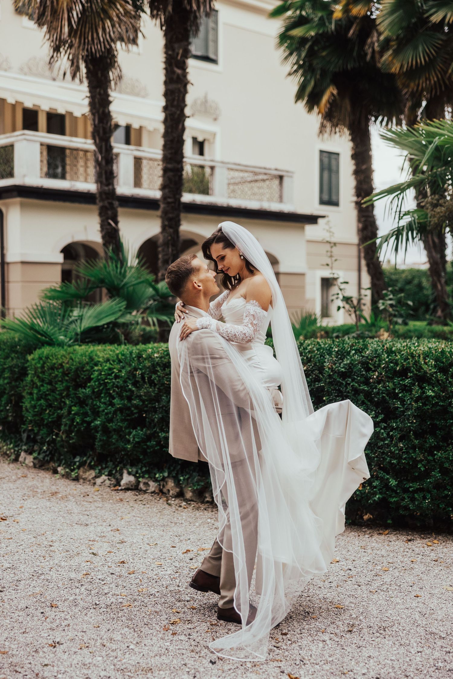 Groom lifts bride in white gown and veil outside elegant villa surrounded by palm trees and lush greenery.