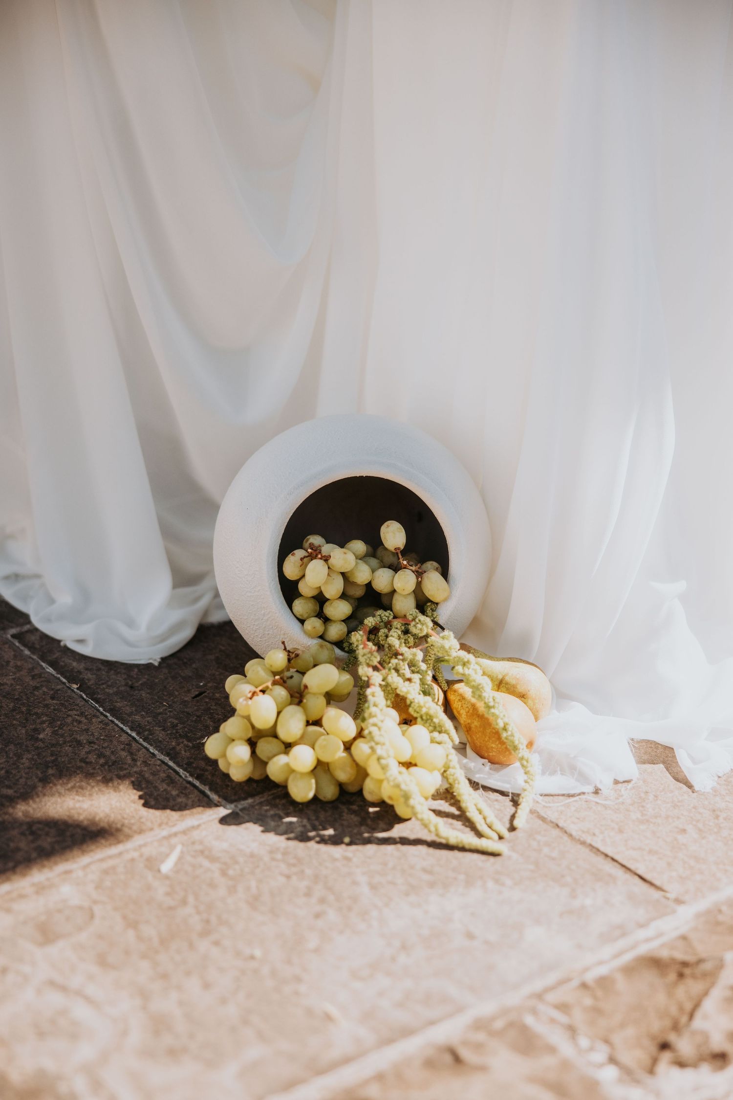 White vase with spilling grapes and fruit decorating an outdoor wedding venue.