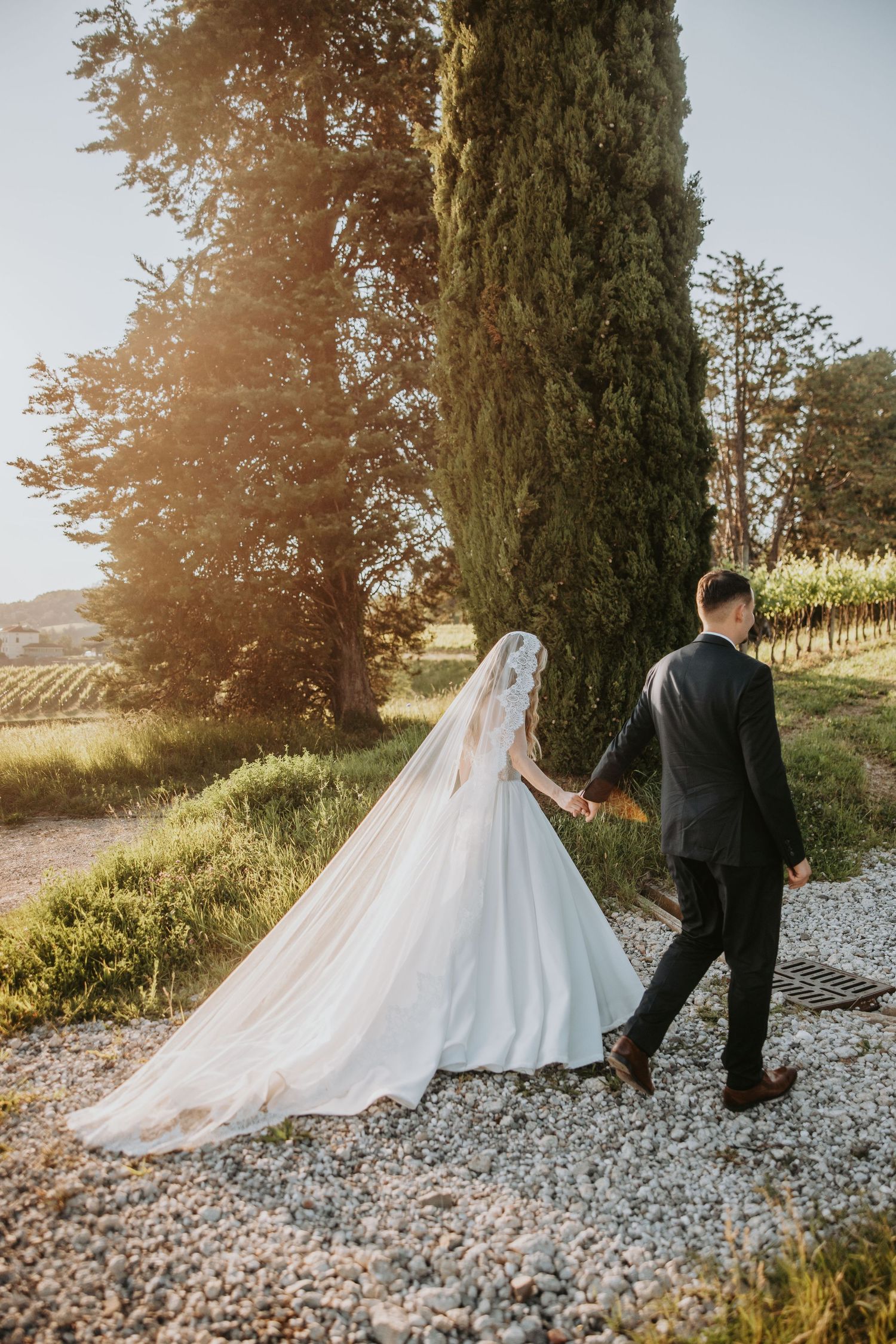 Bride and groom walk hand in hand along a gravel path beside cypress trees at golden hour.