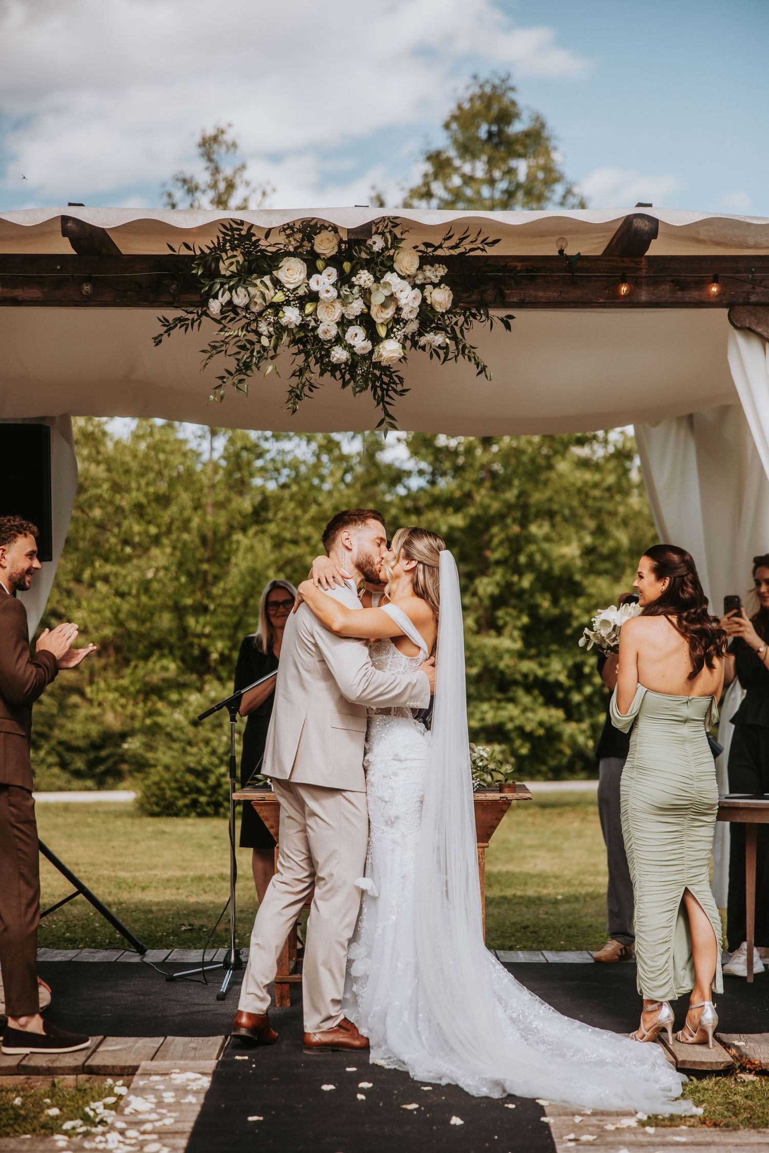 Newlyweds share first kiss at outdoor wedding altar adorned with white floral arrangements.
