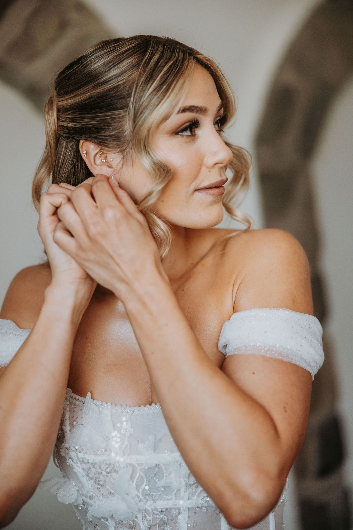 Close-up of blonde bride in off-shoulder lace gown putting on earrings with soft natural lighting.