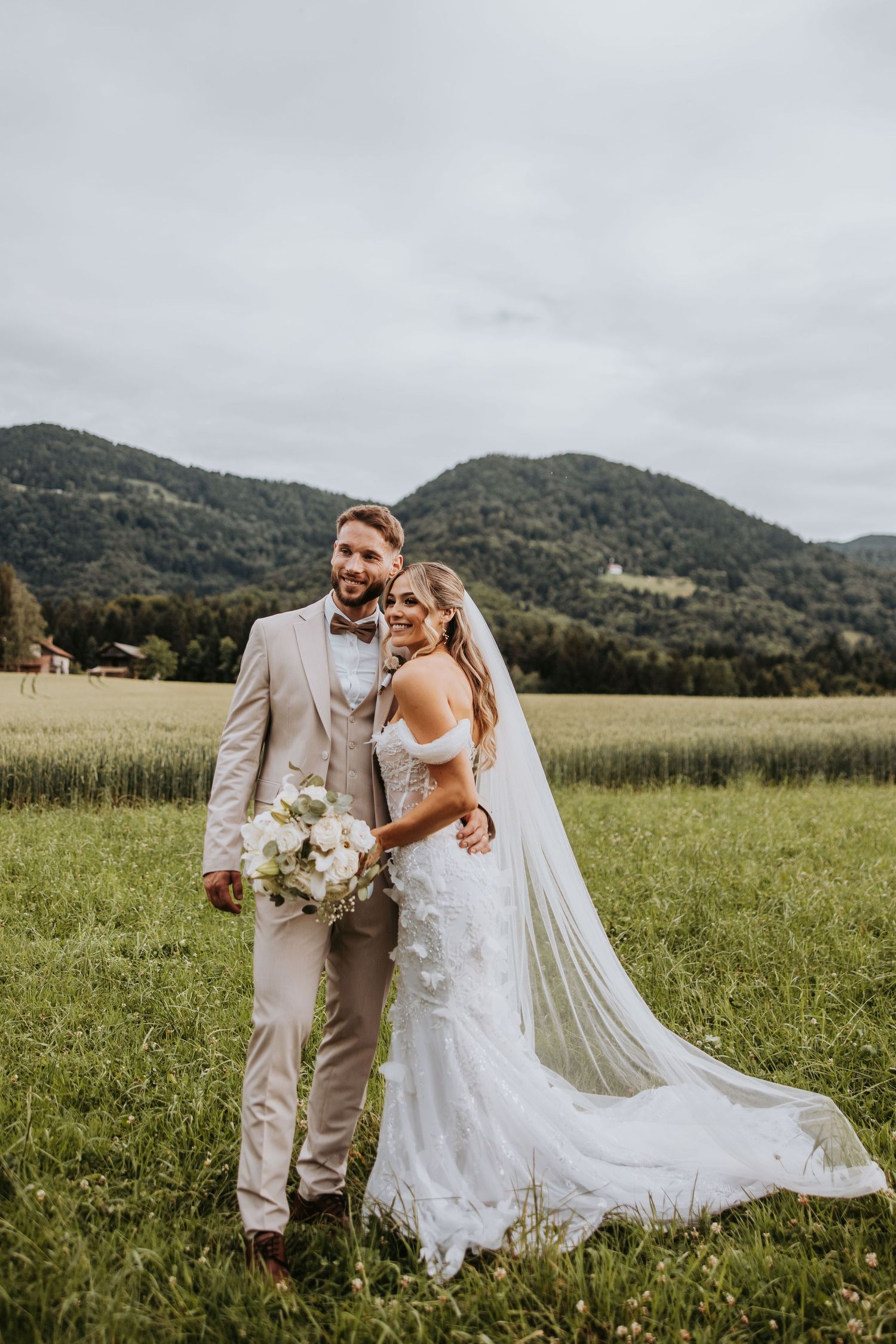 Smiling bride and groom pose in green meadow with stunning mountain backdrop and flowing cathedral veil.