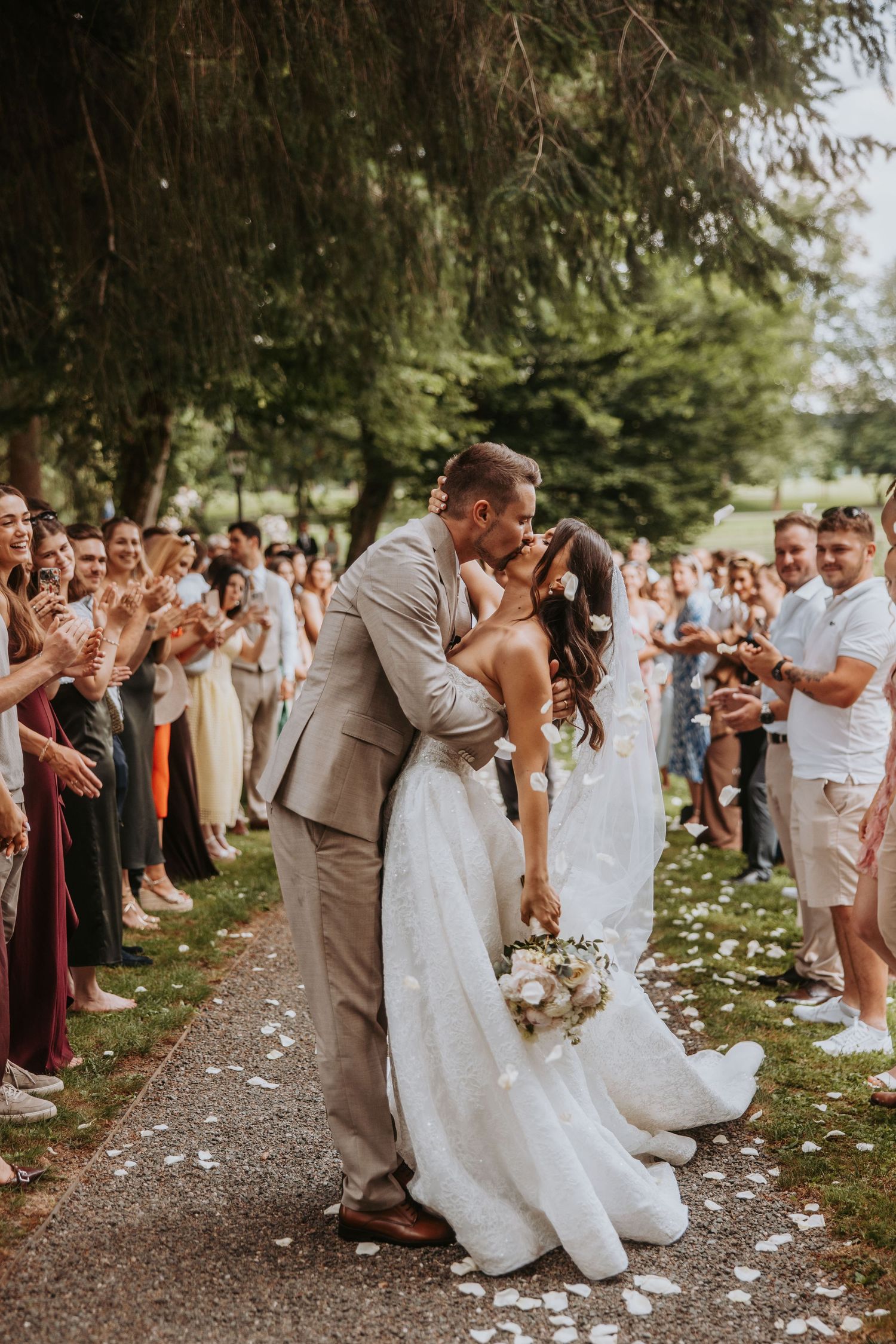 Bride and groom share a romantic kiss while walking down the aisle as wedding guests celebrate with confetti outdoors.