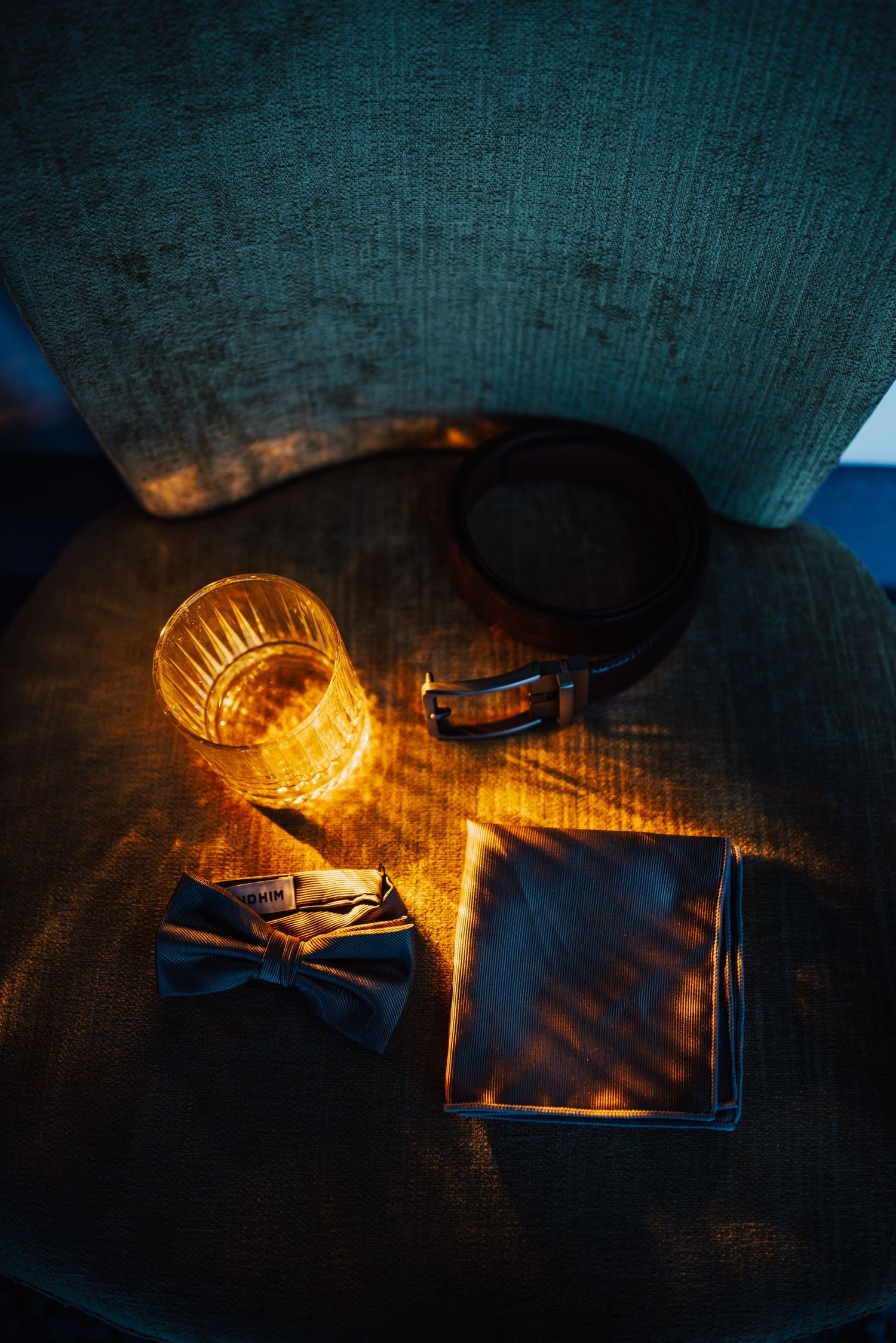 Groom accessories including bow tie, pocket square, leather belt, and whiskey glass in golden light.
