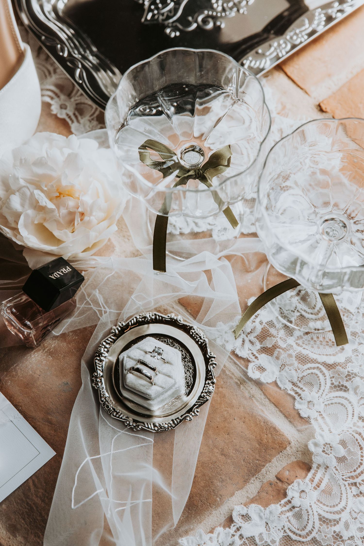 Elegant wedding flat lay with champagne coupes, lace veil, white peony, and diamond ring in ornate silver box.