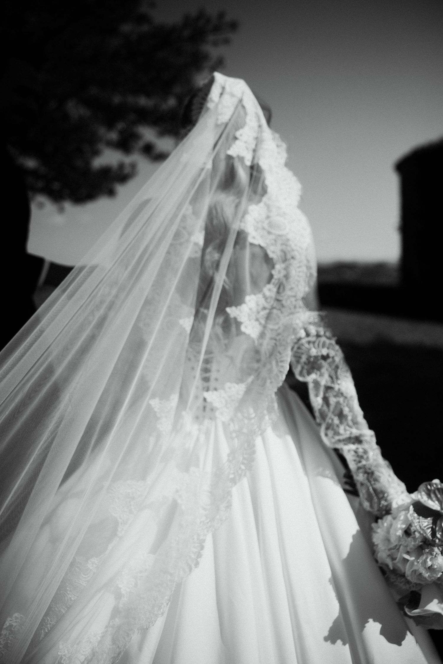 Elegant bride in lace wedding gown and long veil photographed from behind in black and white.