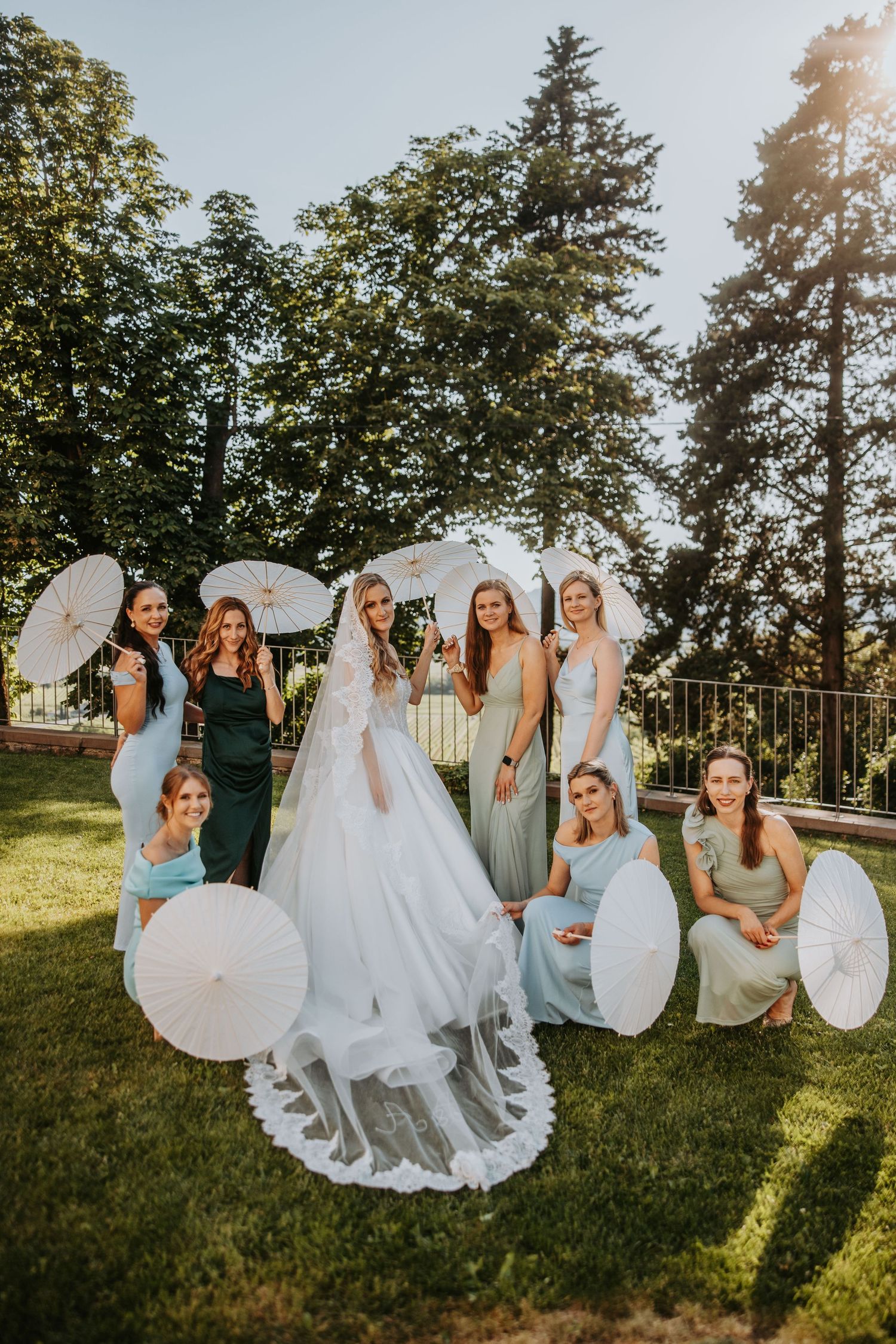 Bride in white gown poses with bridesmaids in mint dresses holding white parasols outdoors.