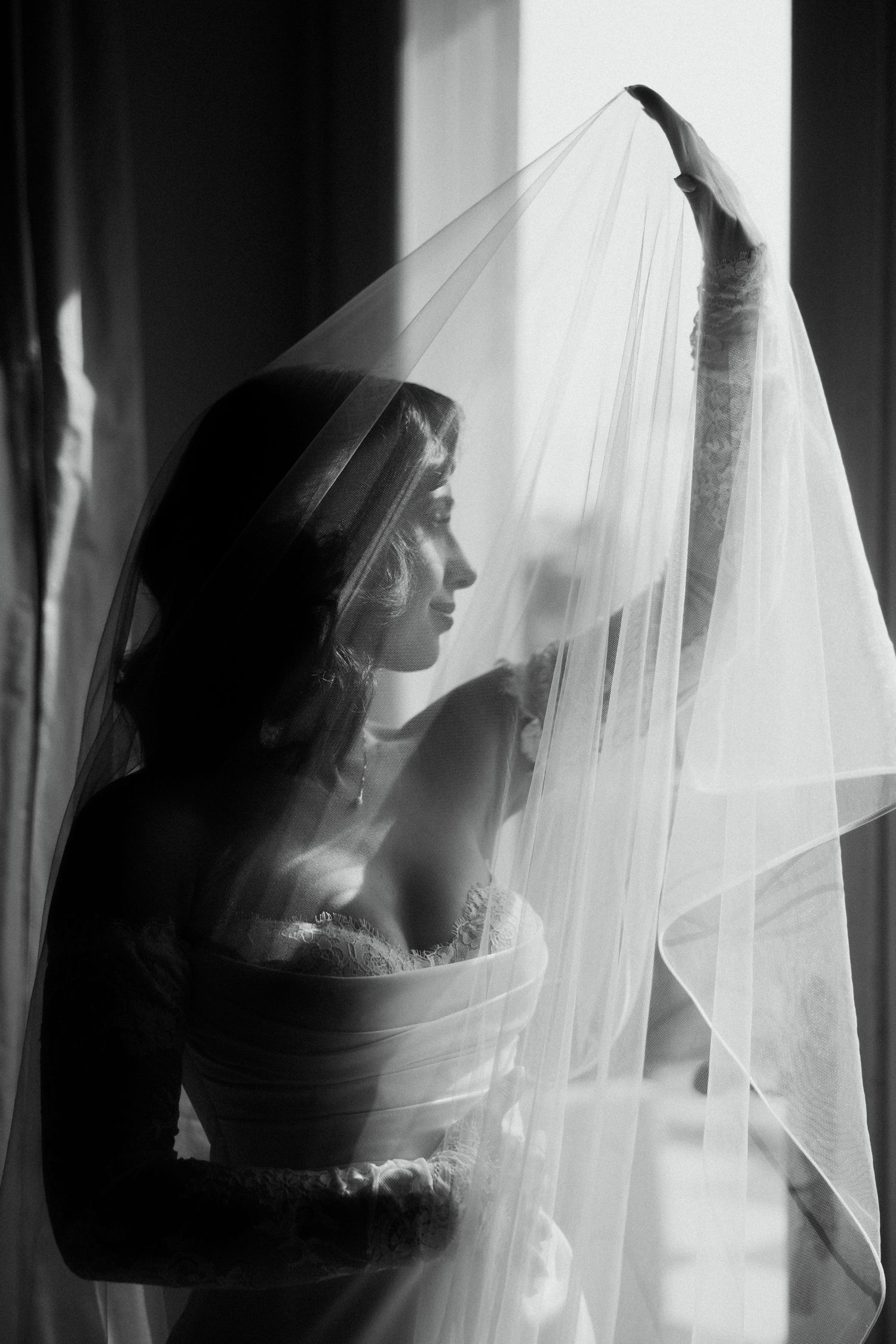 Dramatic black and white portrait of bride holding veil near window in wedding dress.
