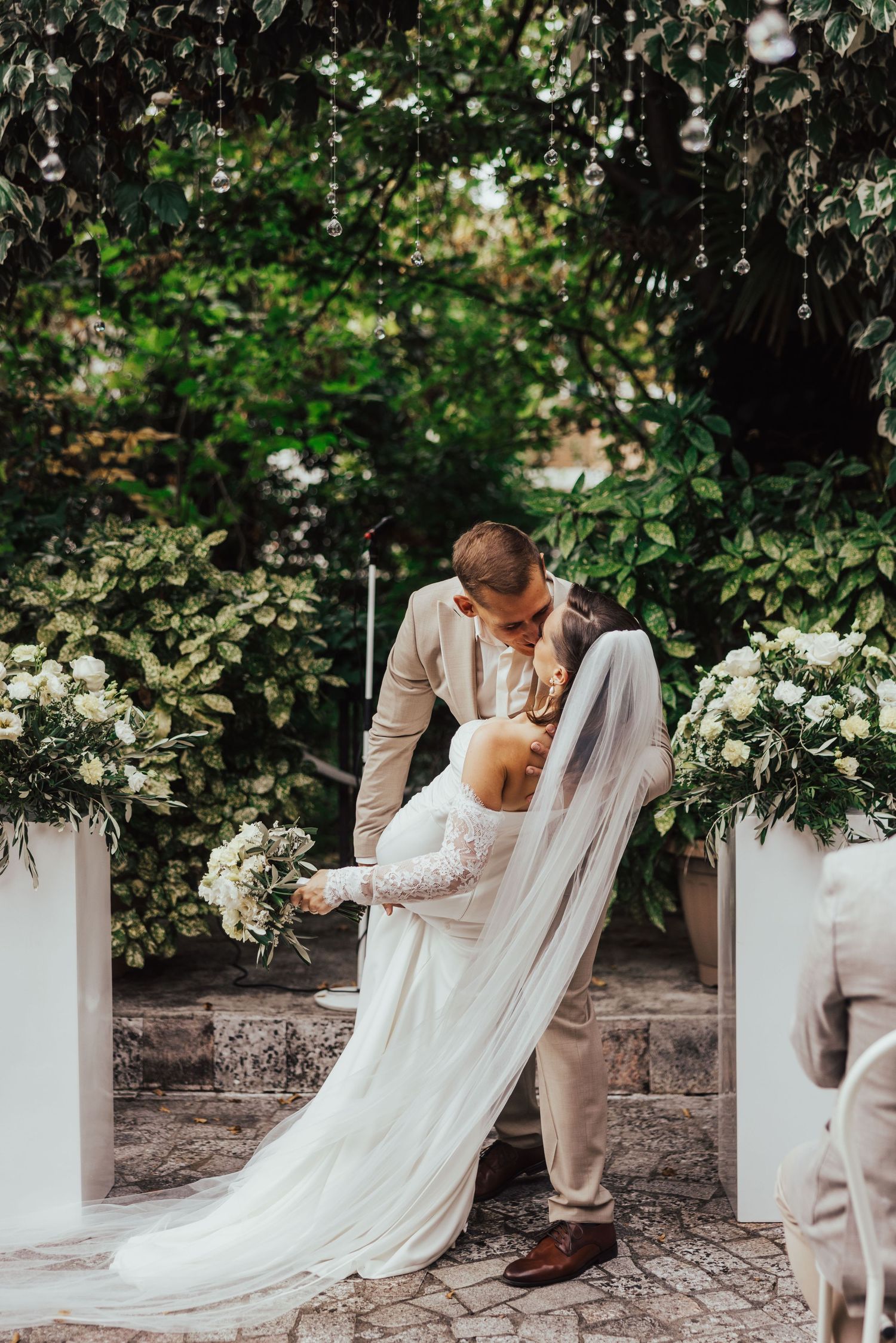 Bride and groom share a romantic dip kiss at an outdoor wedding ceremony surrounded by lush greenery and white floral arrangements.