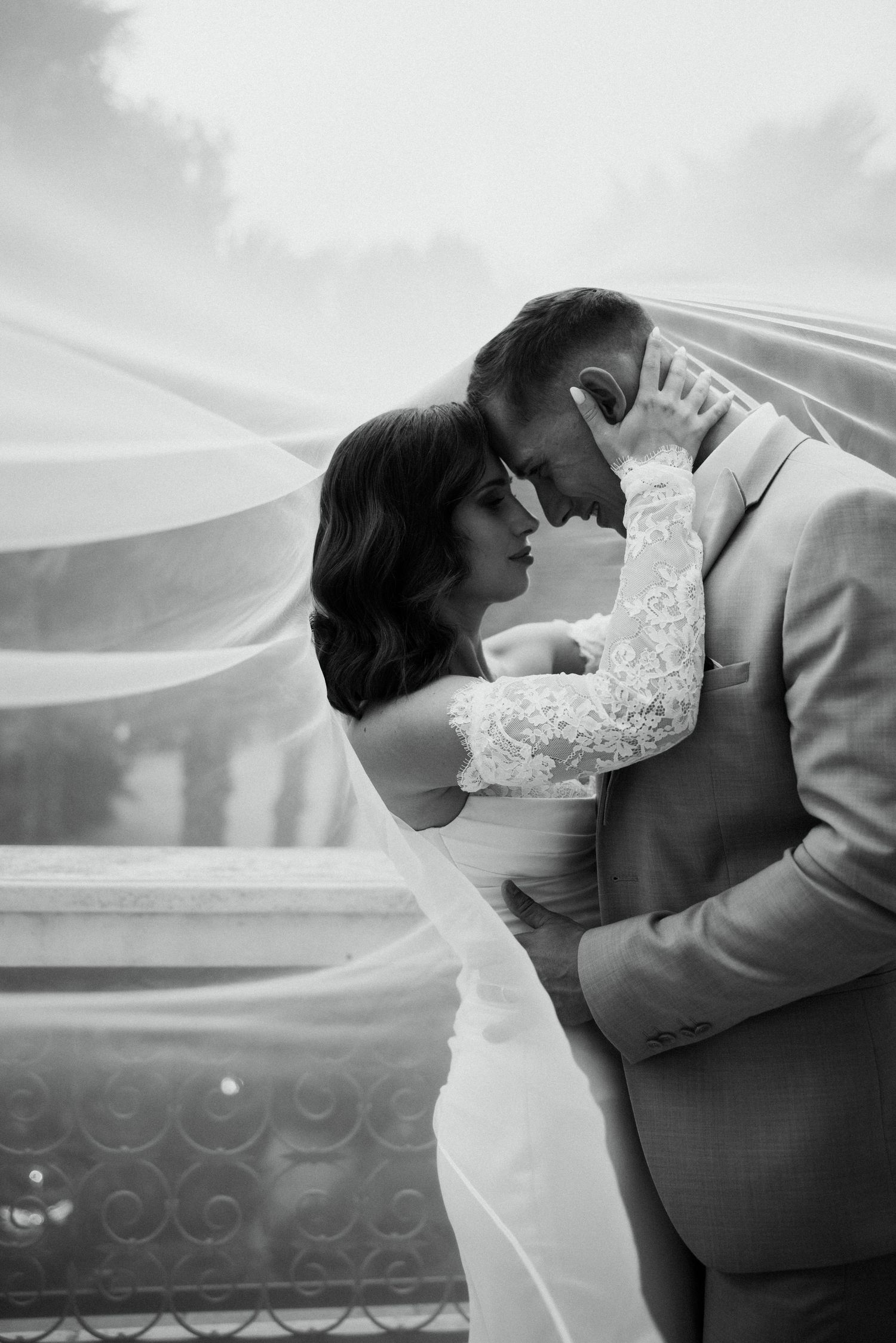 Romantic black and white photo of bride and groom embracing under flowing veil outdoors.
